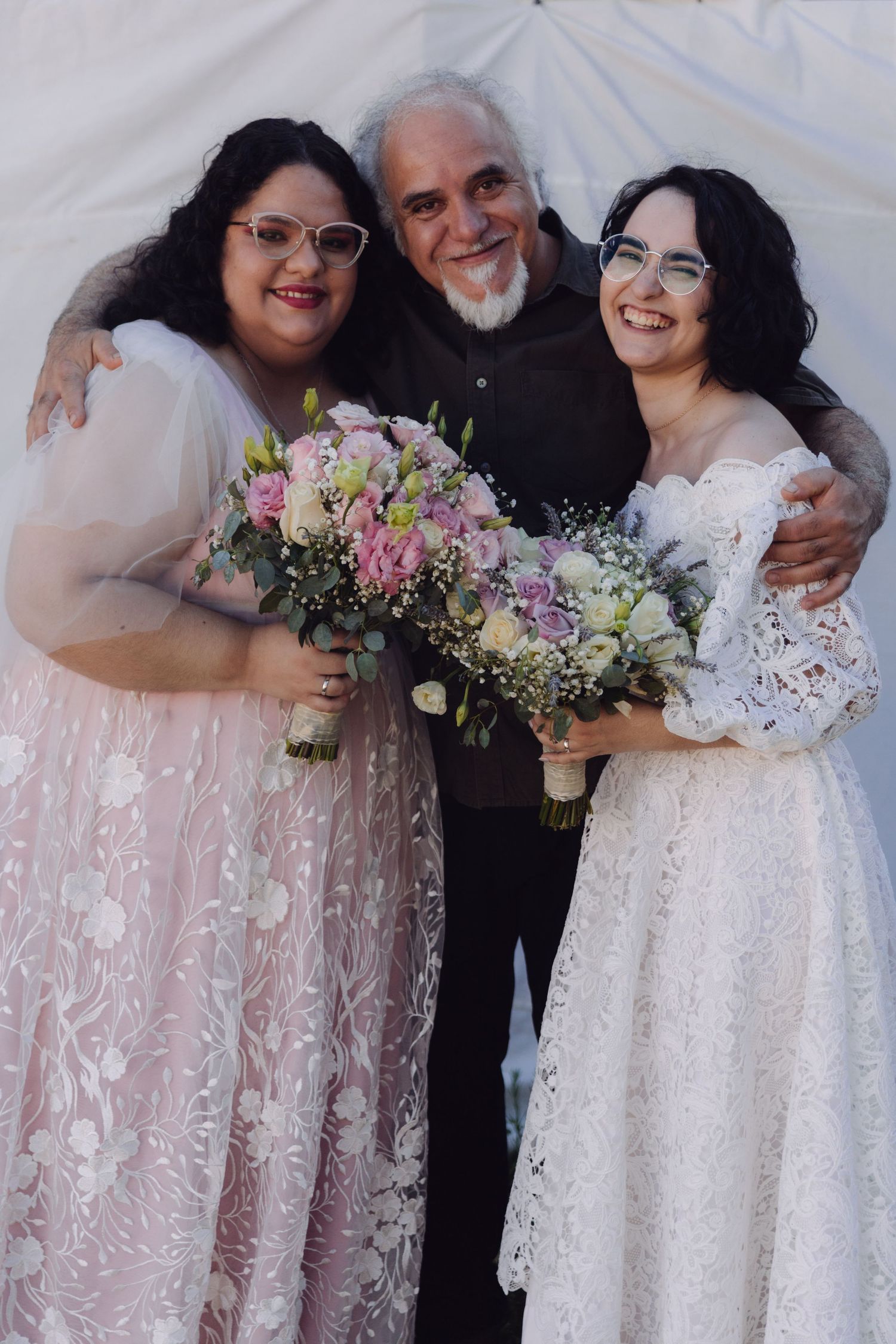 Two brides holding floral bouquets smile with a bearded man at a wedding celebration.