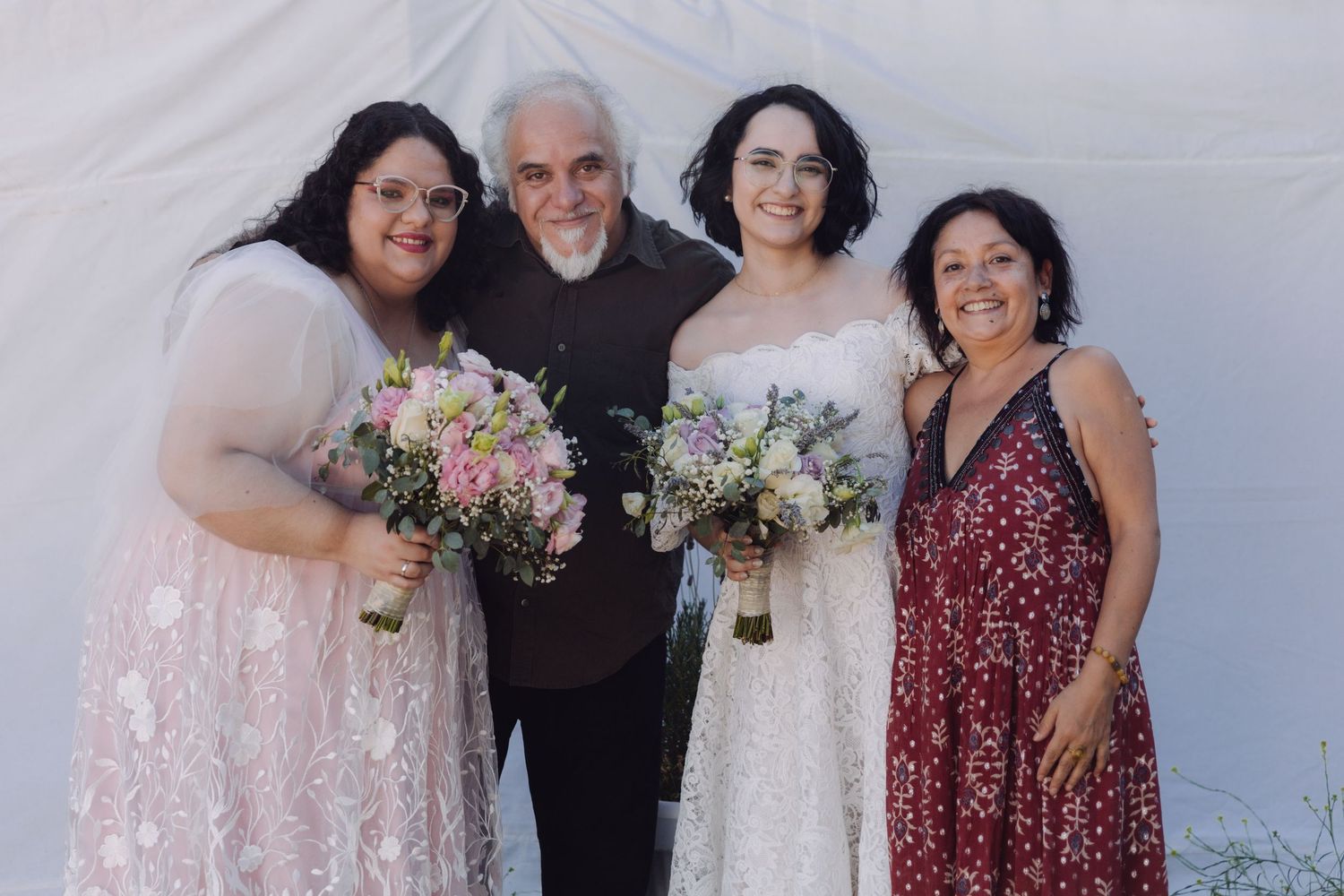 Two brides with bouquets pose with an older man and woman at a wedding celebration.
