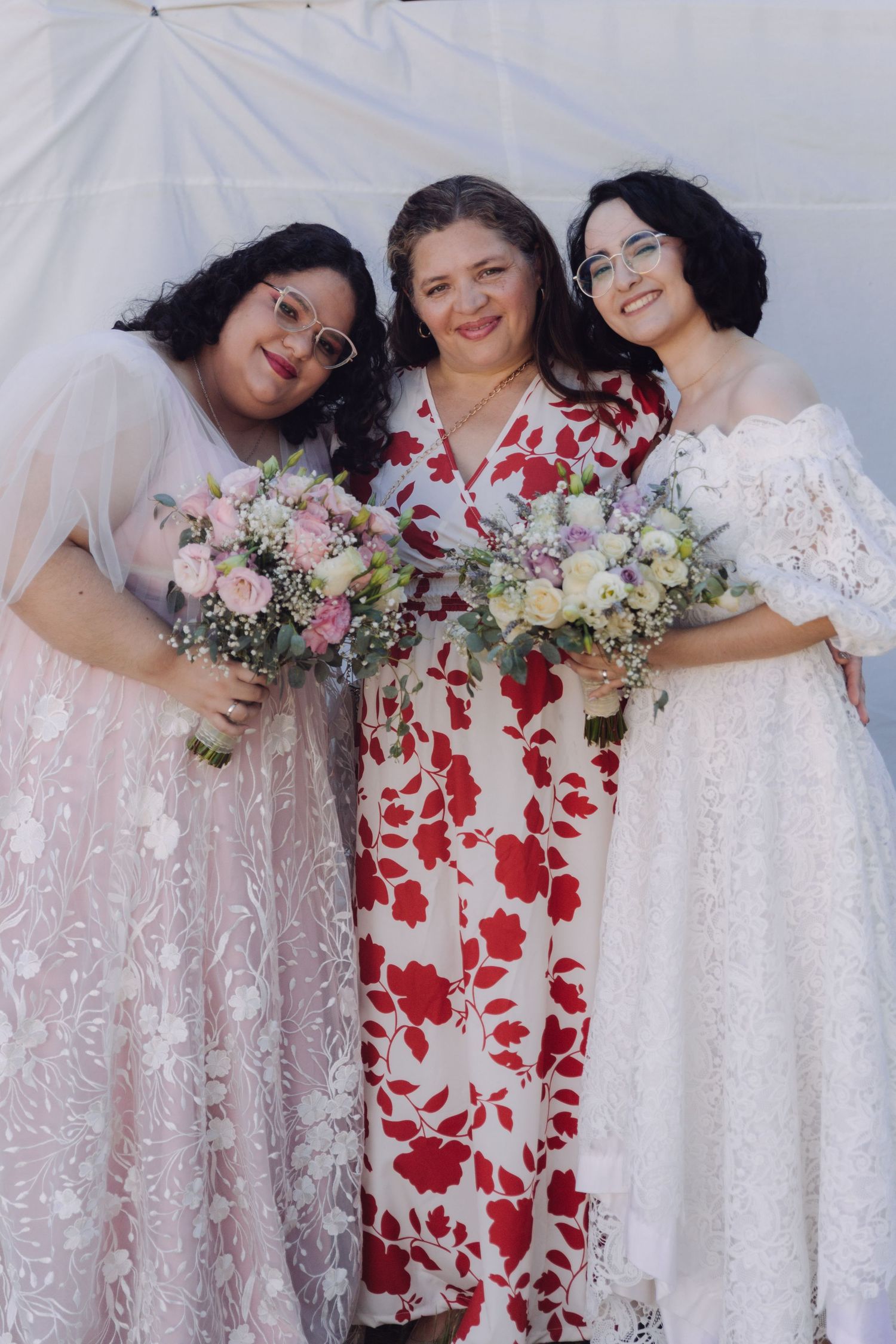 Two brides in lace gowns holding bouquets pose with a smiling woman in a floral dress.