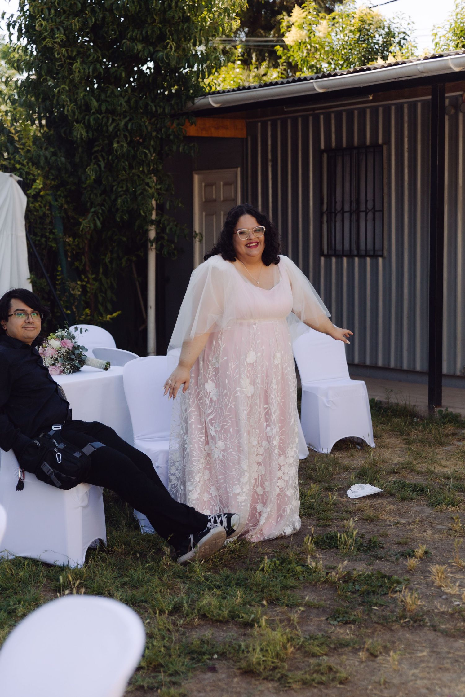 A bride in a pink lace gown poses outdoors at a wedding venue with white chairs.