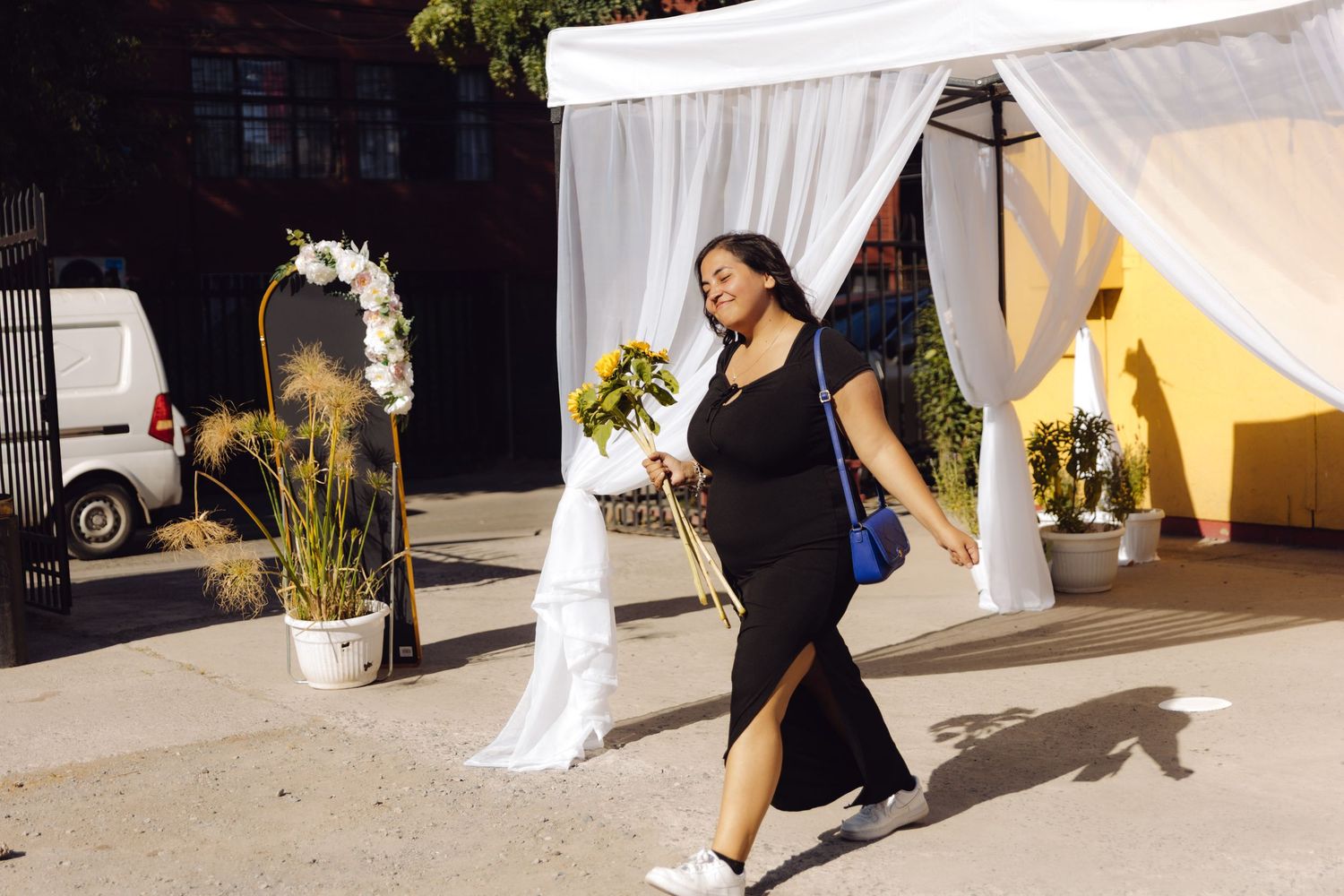 Woman in black dress carrying blue purse and sunflowers walks past white draped wedding arch outdoors.