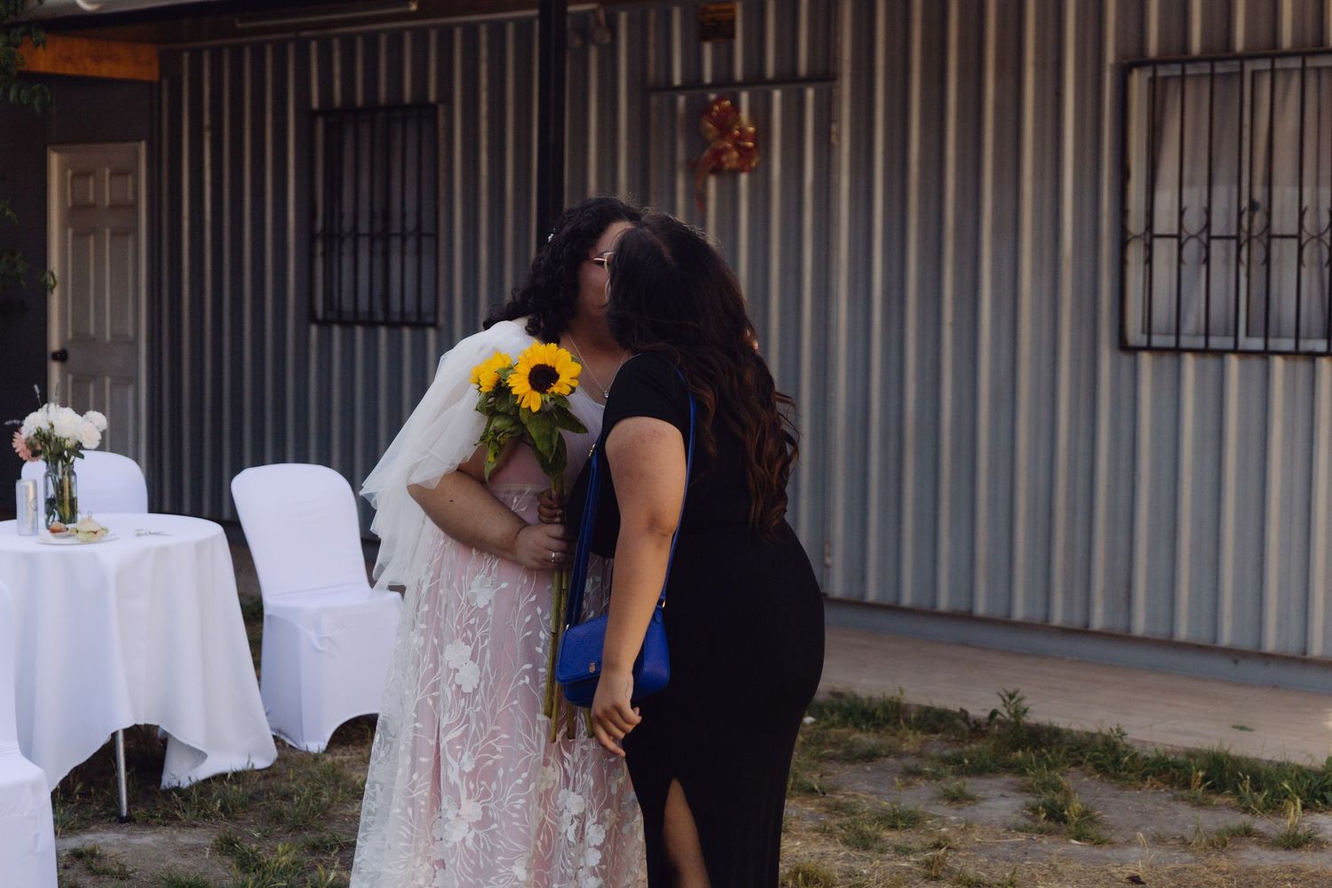 Bride in lace wedding gown embraces guest while holding sunflower bouquet at outdoor wedding.