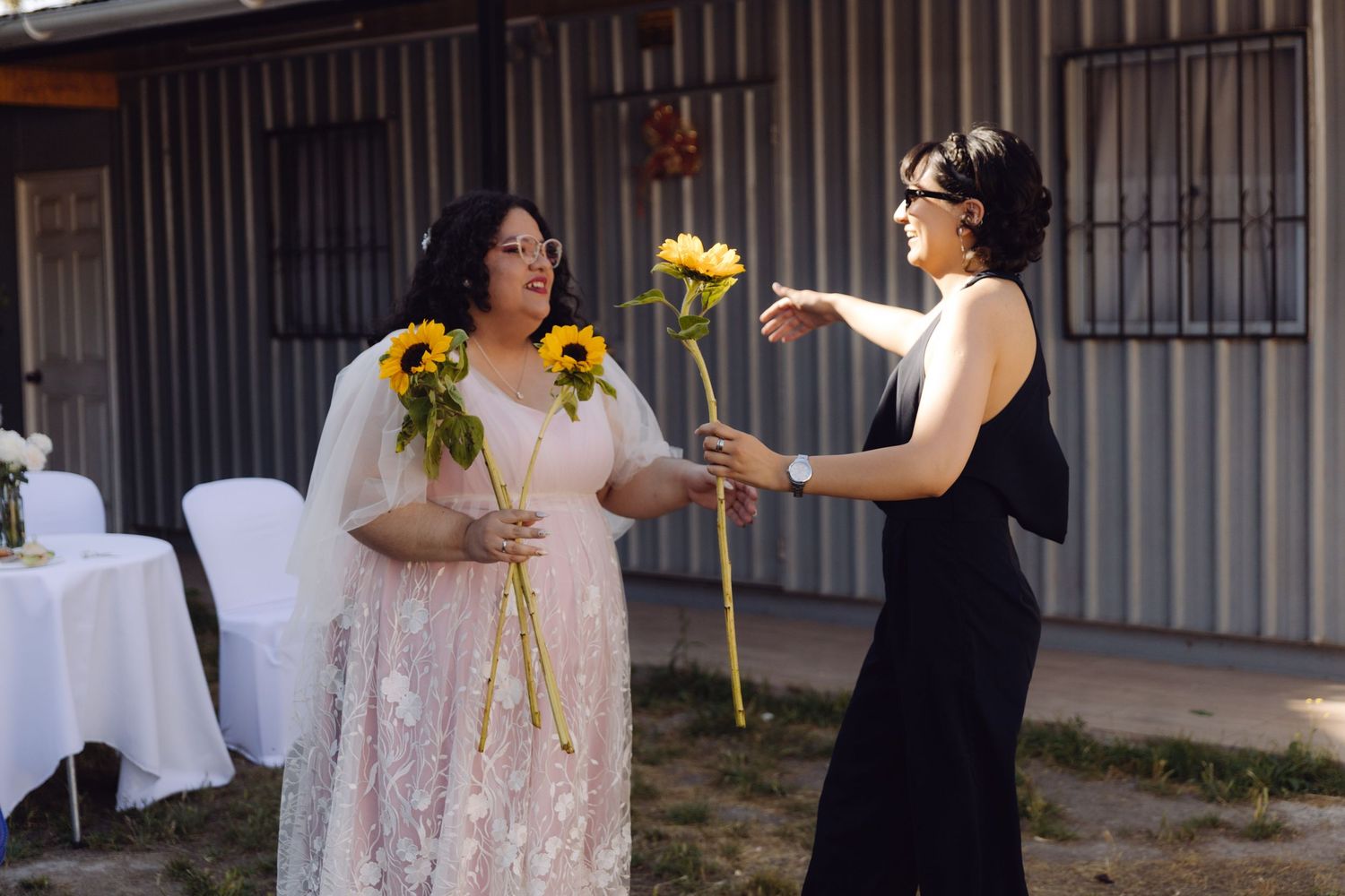 Bride in pink lace gown receives sunflowers from woman in black dress at outdoor wedding venue.