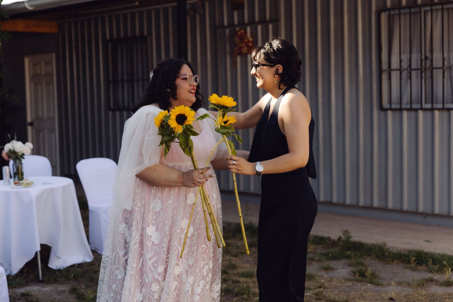 Bride in floral lace dress smiles as guest in black dress hands her sunflowers at outdoor wedding.