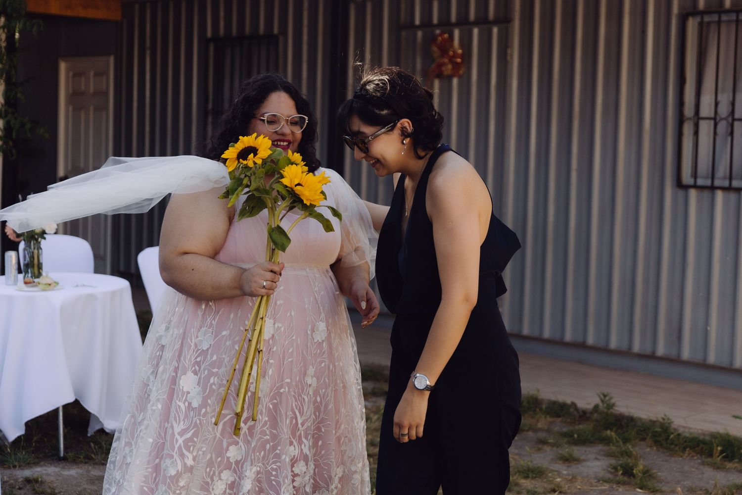 Bride in pink lace gown and veil examines sunflower bouquet with guest in black outfit outdoors.