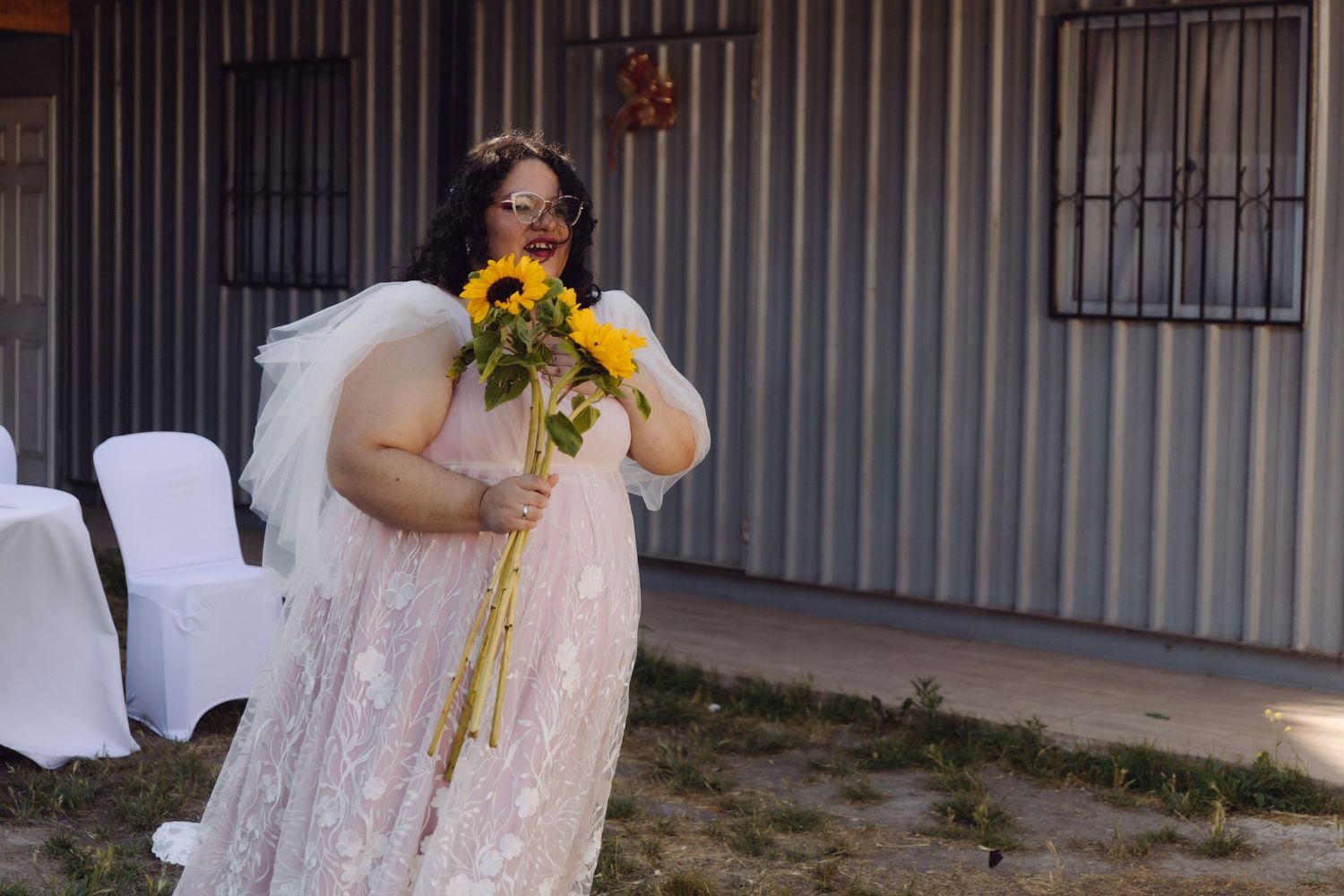 Plus-size bride in pink lace gown holds large sunflower bouquet near corrugated metal building.