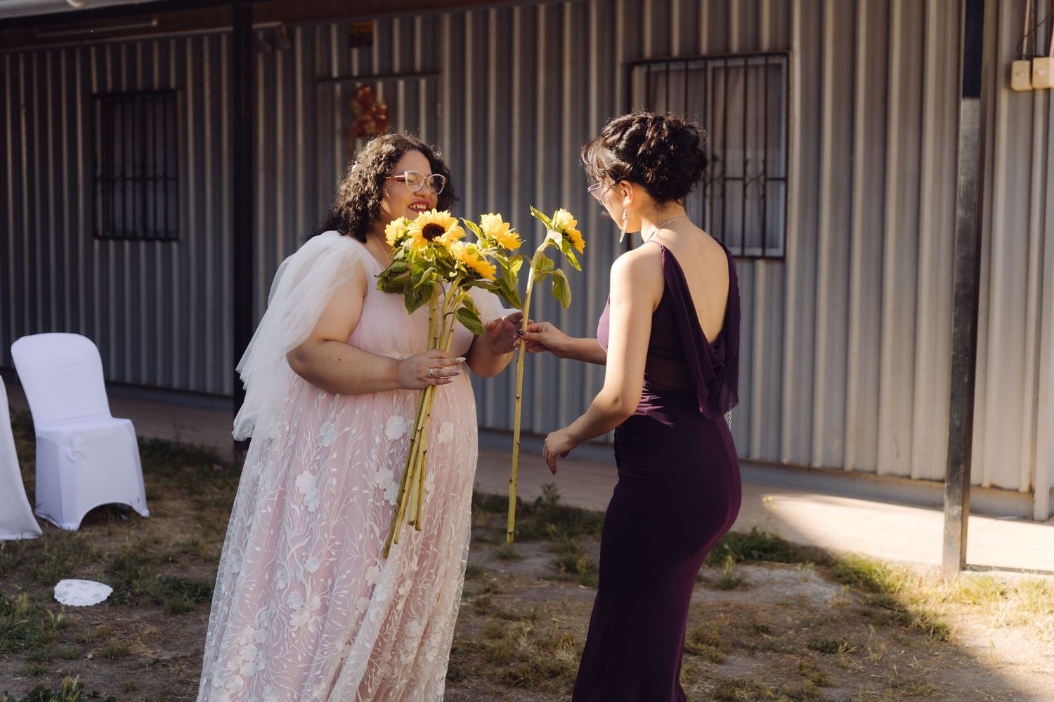 Bride in lace gown and veil receives sunflowers from guest in dark dress at outdoor wedding.