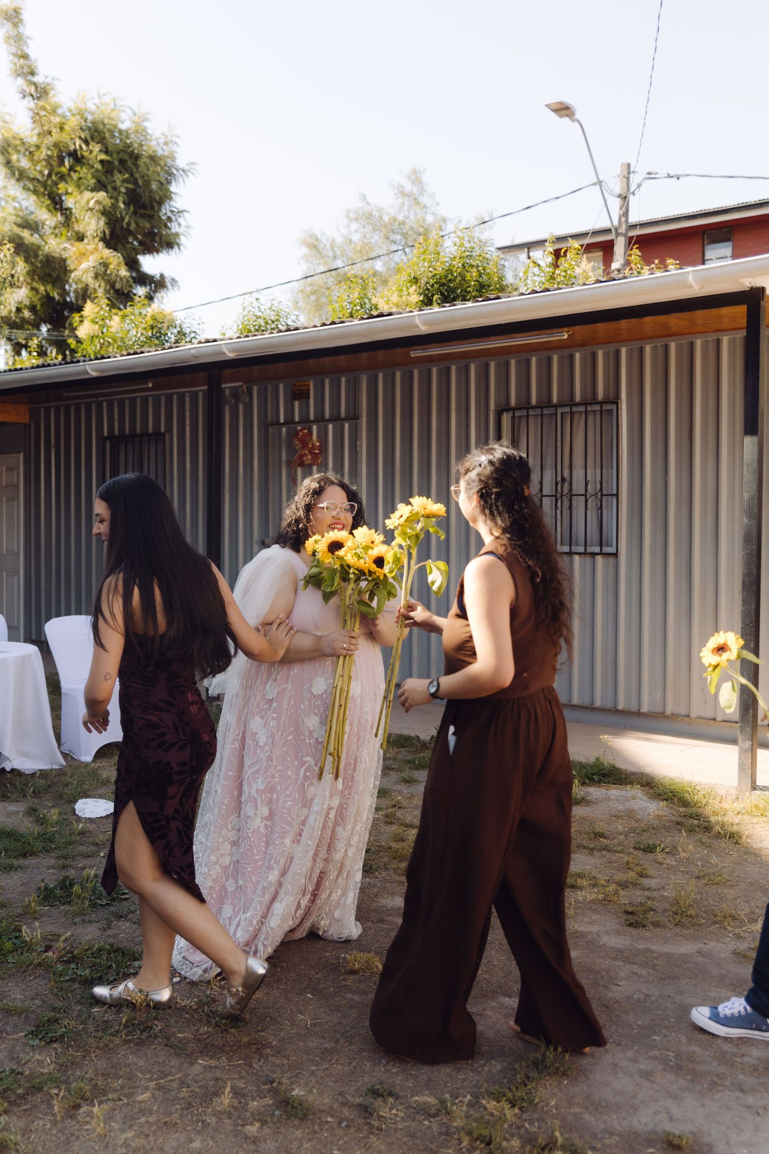 Bride in pink lace gown accepts sunflower bouquet from guests in brown dresses at outdoor wedding.