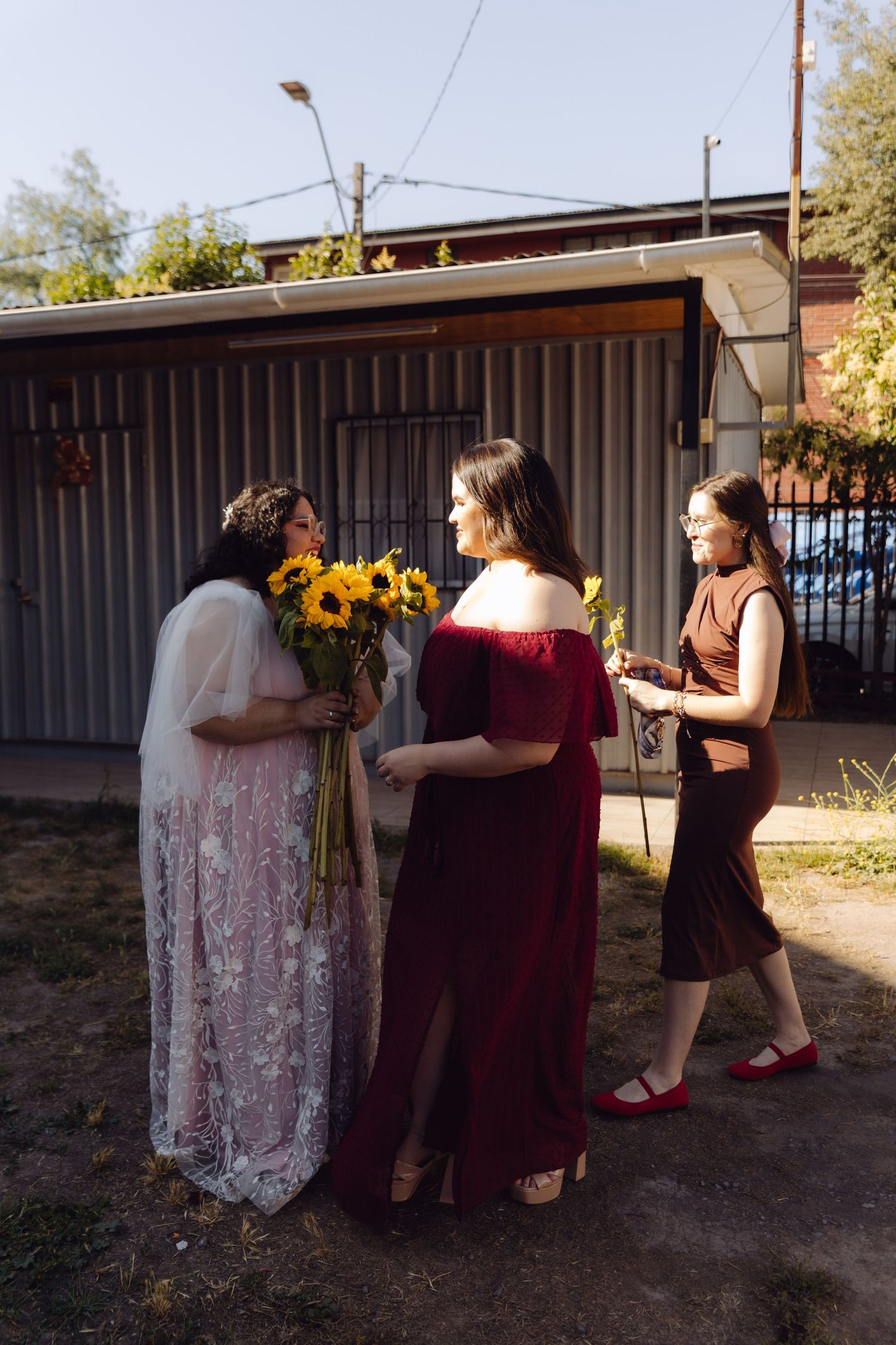 Bride holding sunflowers chats with guests in burgundy and brown dresses at outdoor wedding.