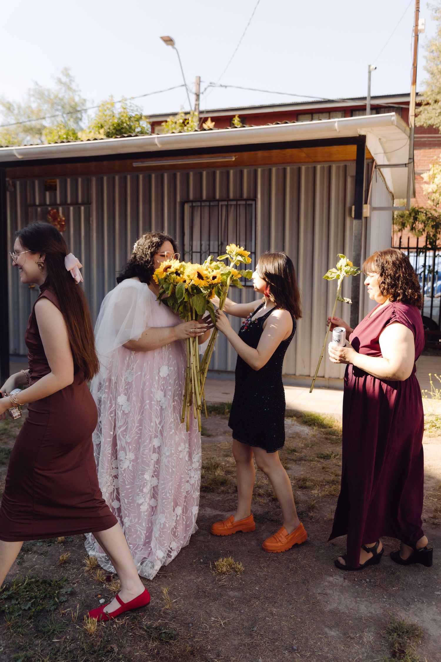 Bride in lace gown receives sunflower bouquet from woman in black dress among wedding guests outdoors.