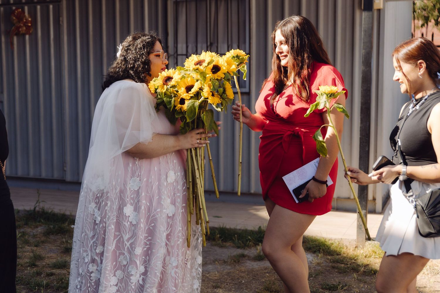 Bride in lace gown receives sunflower bouquet from woman in red dress at outdoor wedding celebration.