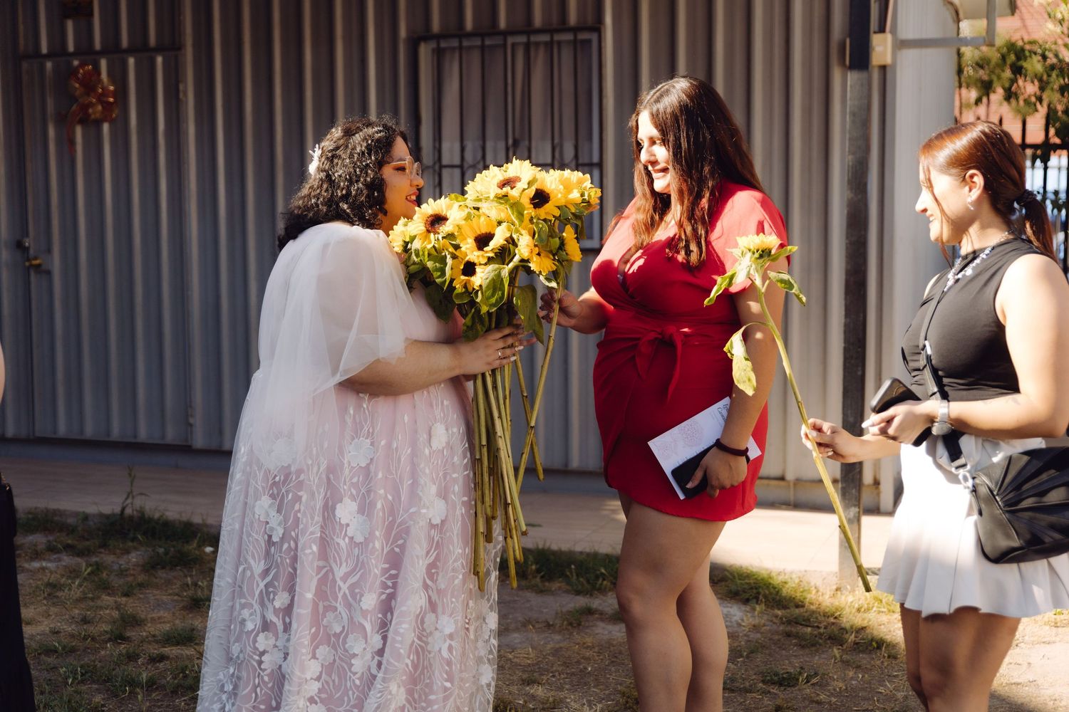 Wedding guests exchange vibrant sunflower bouquets outside a rustic venue on a sunny day.