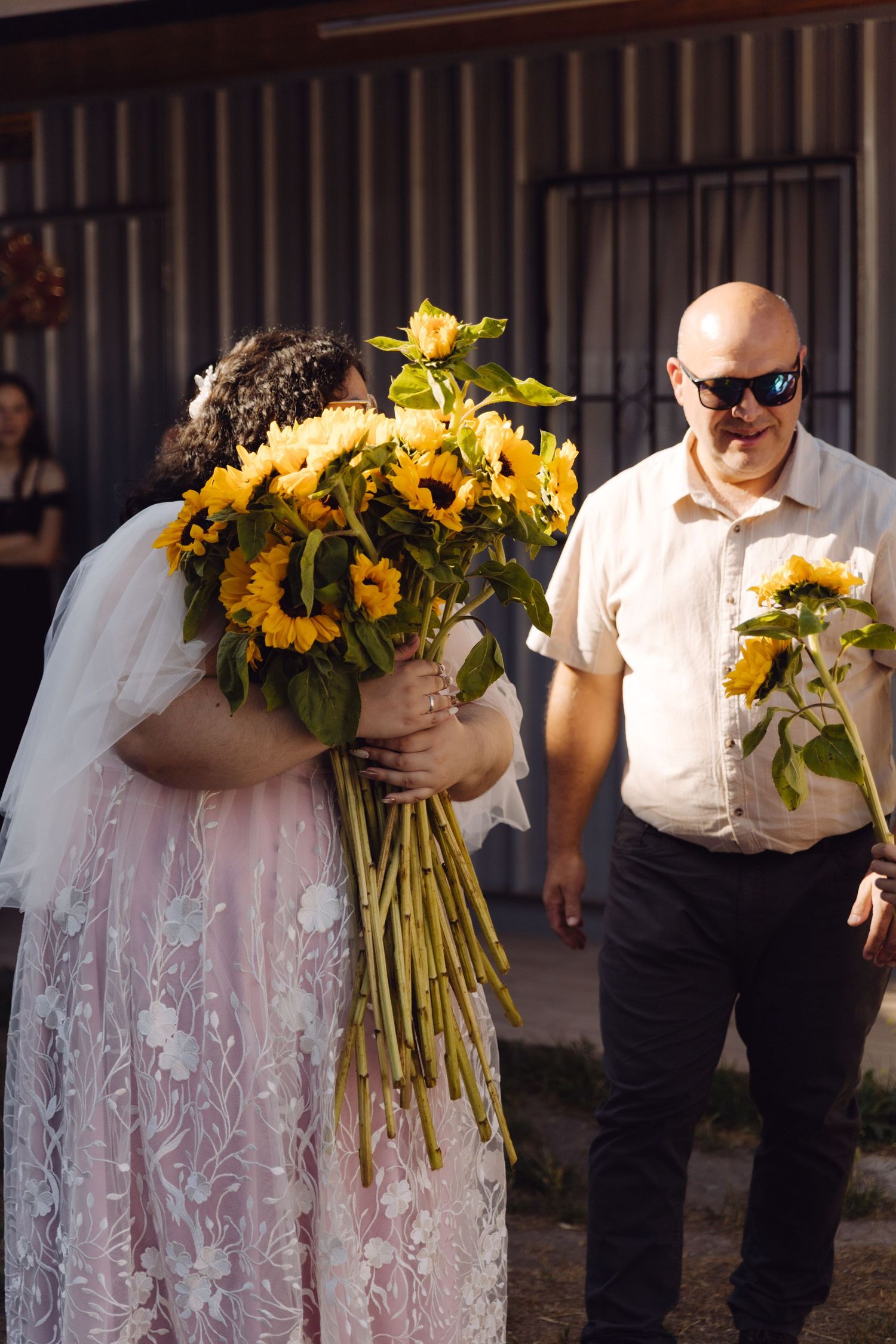 Bride in pink lace gown buries face in large sunflower bouquet as bald man smiles behind her.
