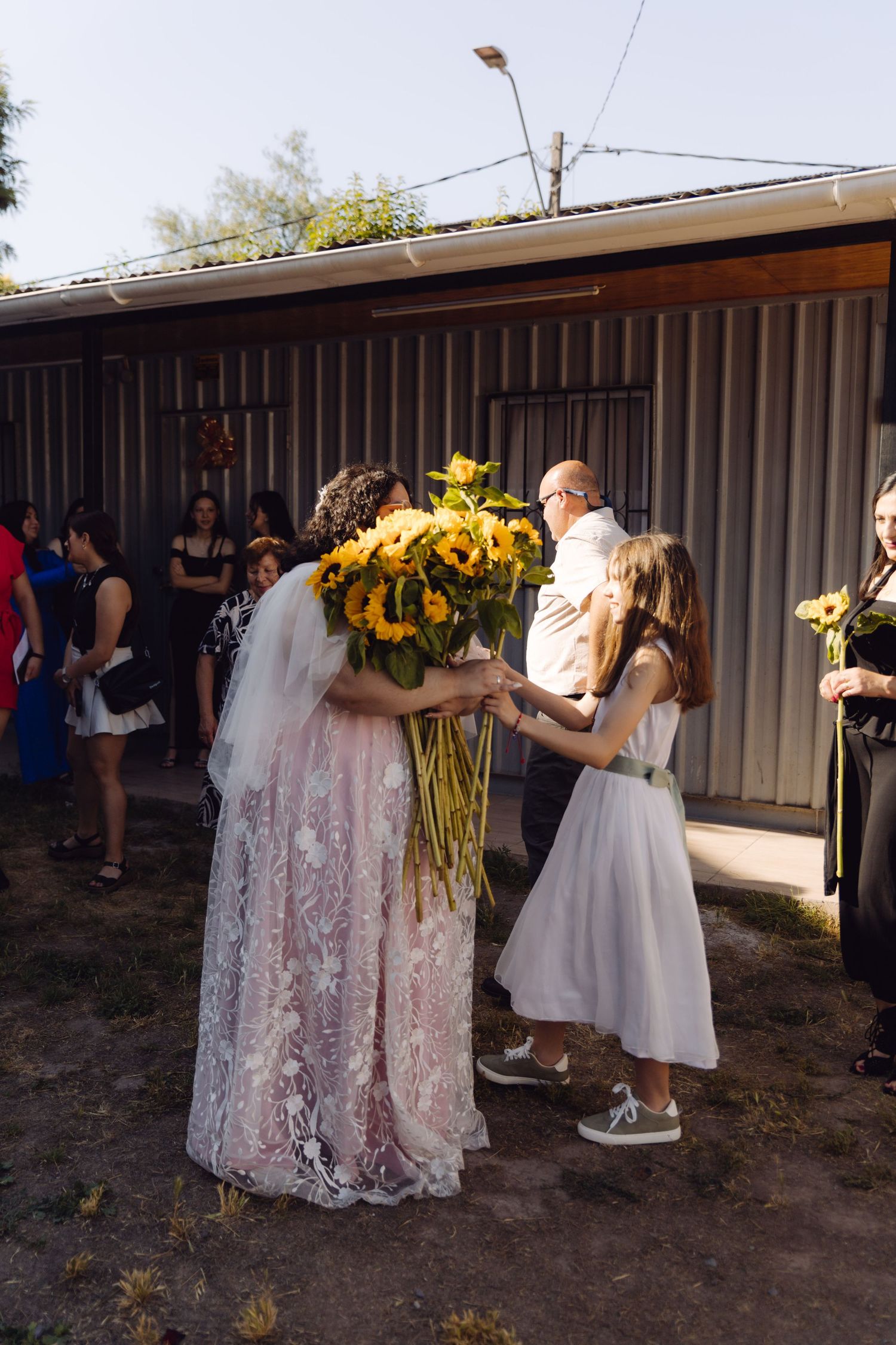 Bride in lace veil accepts sunflowers from young girl in white dress at outdoor wedding.