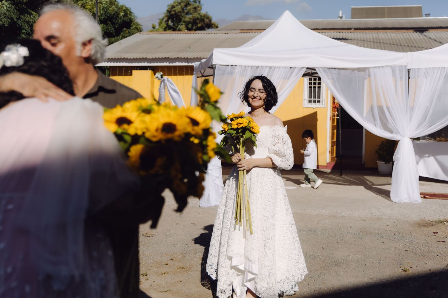 Smiling bride in white lace dress holds sunflowers near decorated canopy at outdoor wedding.