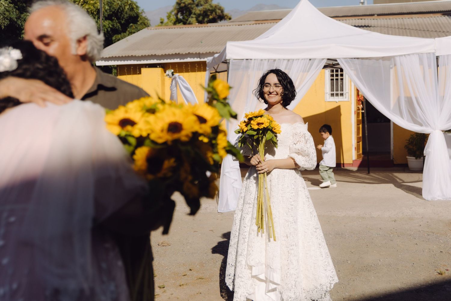 Radiant bride in white lace gown smiles while holding sunflower bouquet at sunny outdoor wedding.