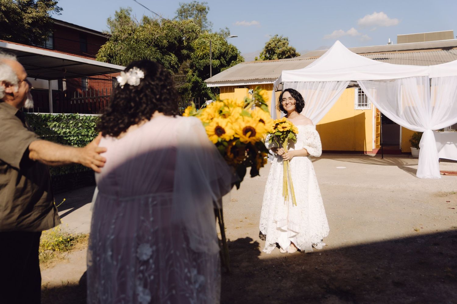 Two brides in white dresses share a joyful sunflower bouquet moment at outdoor wedding venue.