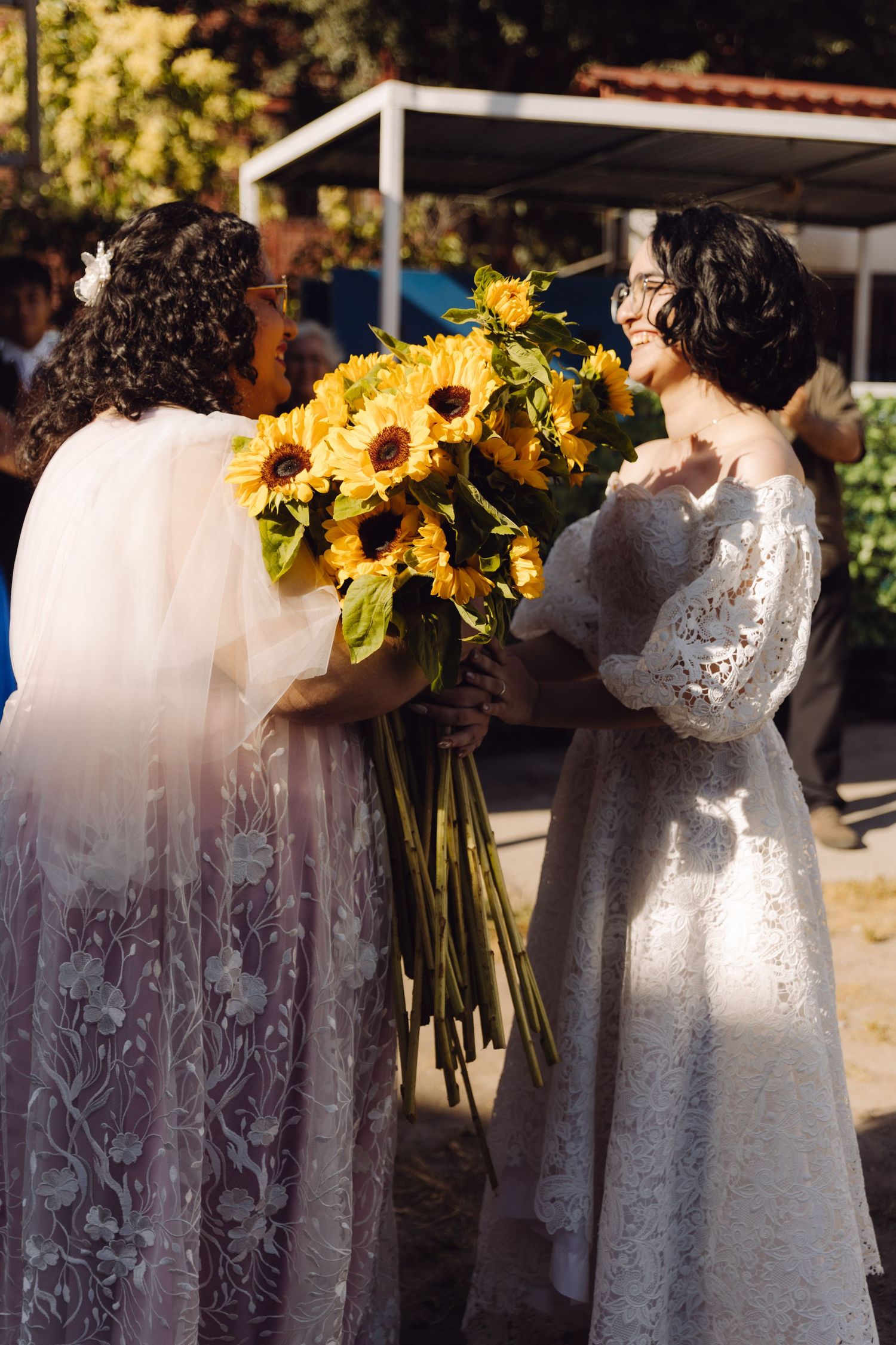 Two brides in white lace gowns share a joyful moment holding large sunflower bouquets outdoors.