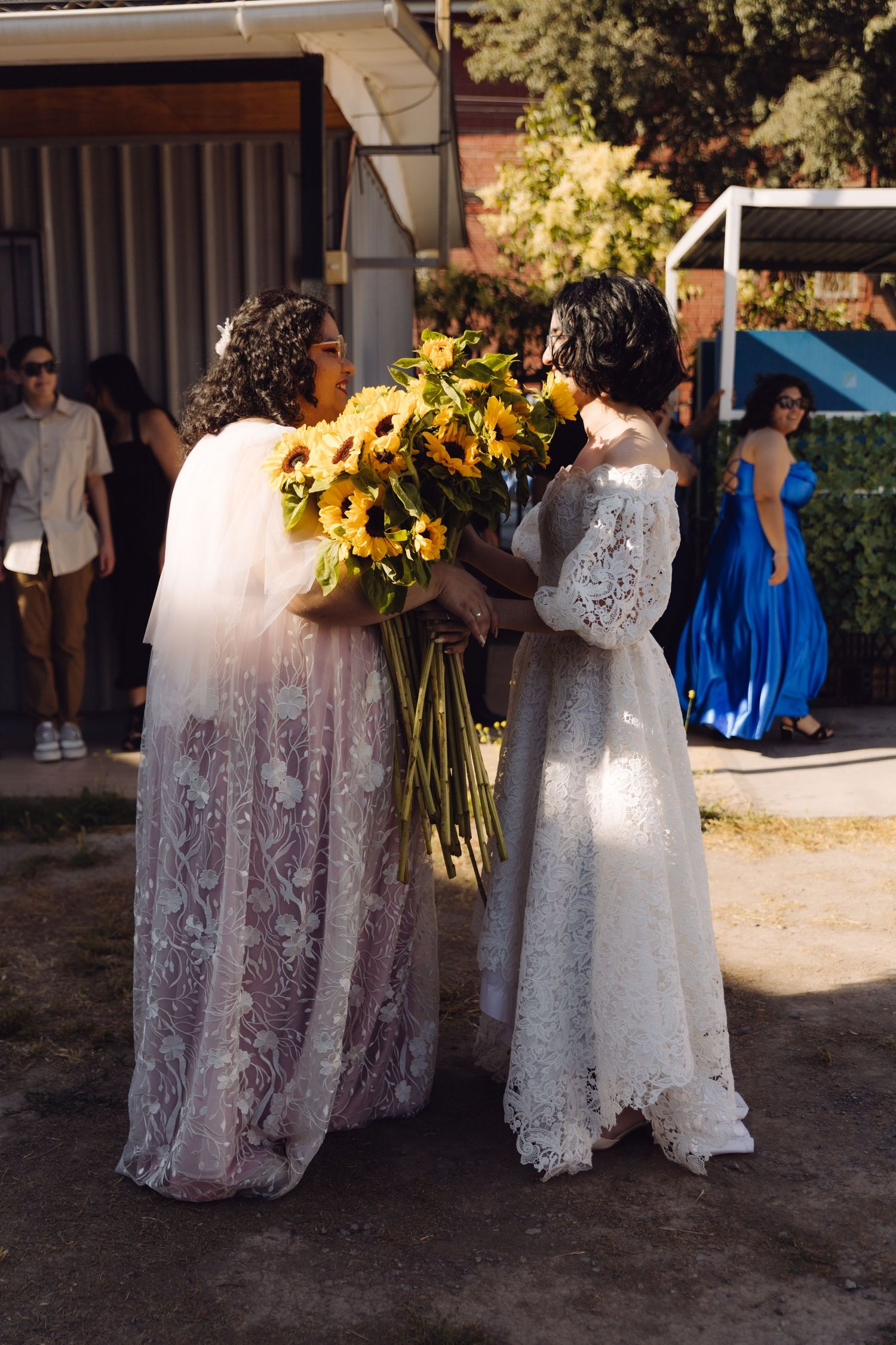 Two brides in white lace gowns stand together holding sunflowers while guests watch at wedding.