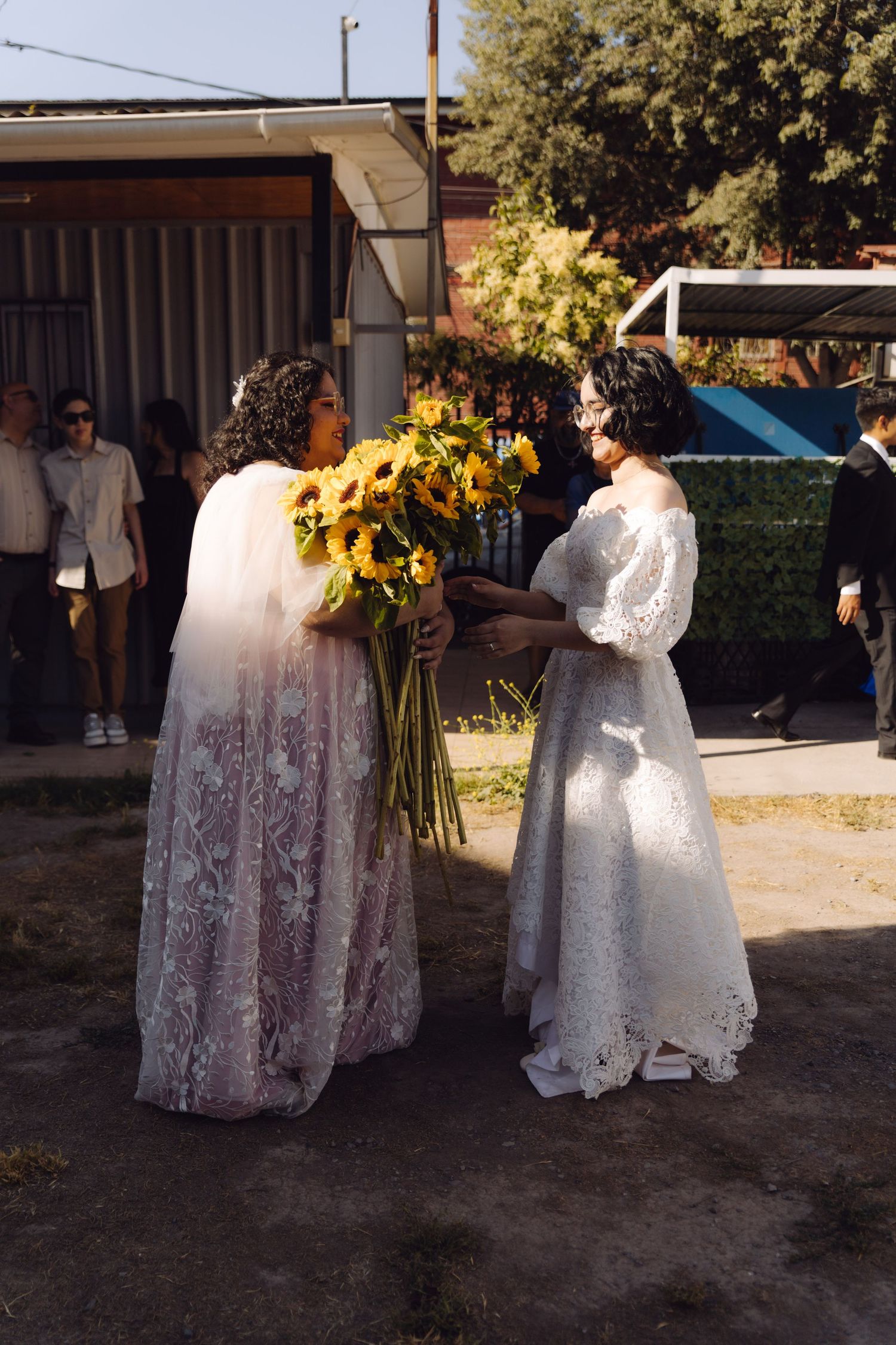 Two brides in lace wedding gowns exchanging sunflower bouquet outdoors at wedding celebration.