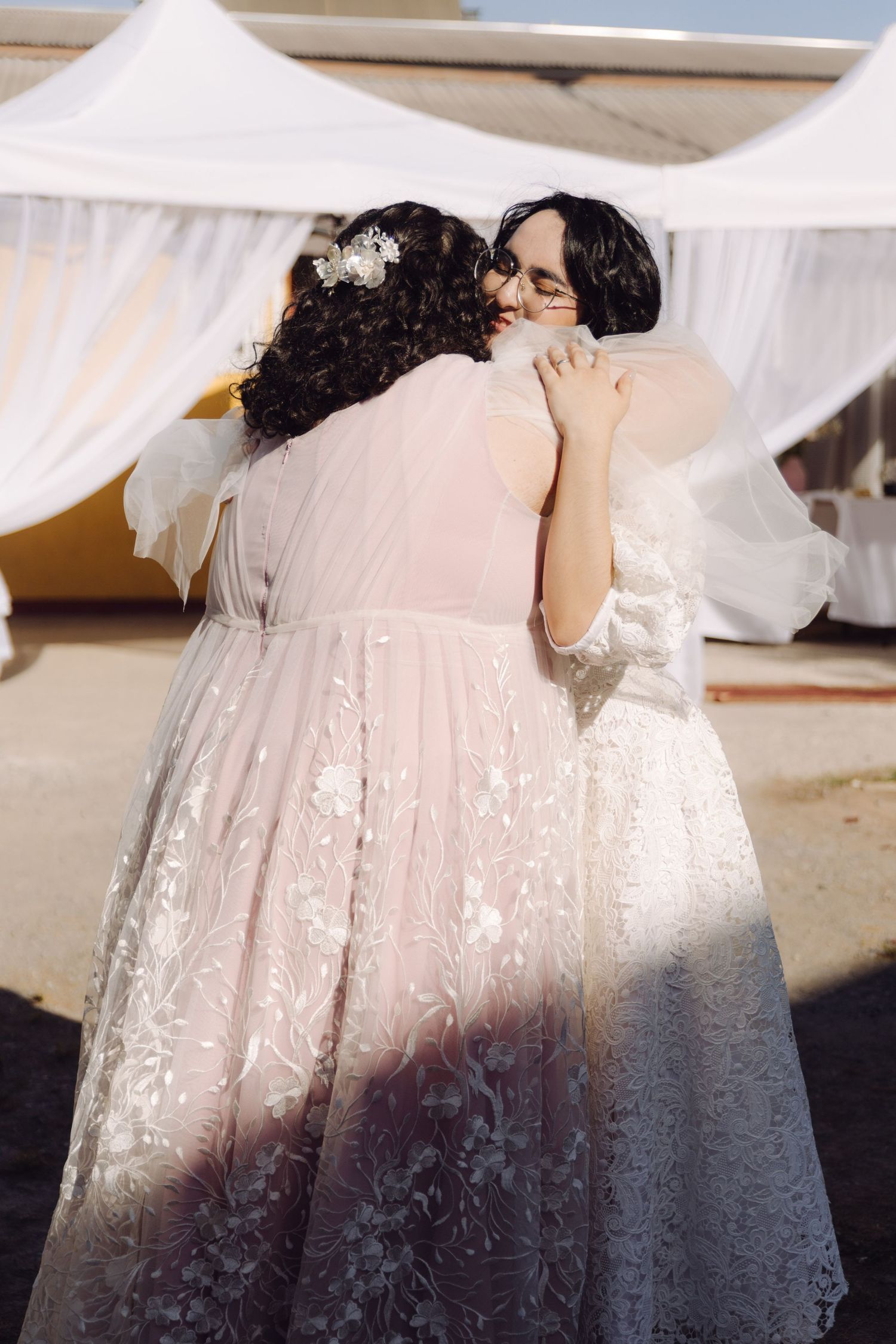 Two brides in white and blush lace gowns sharing tender embrace at outdoor wedding reception.