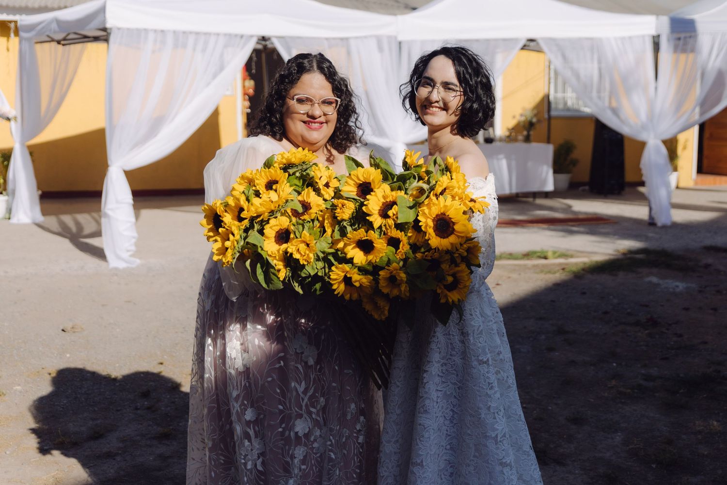 Two women in elegant lace gowns smiling while holding large sunflower bouquet at wedding.