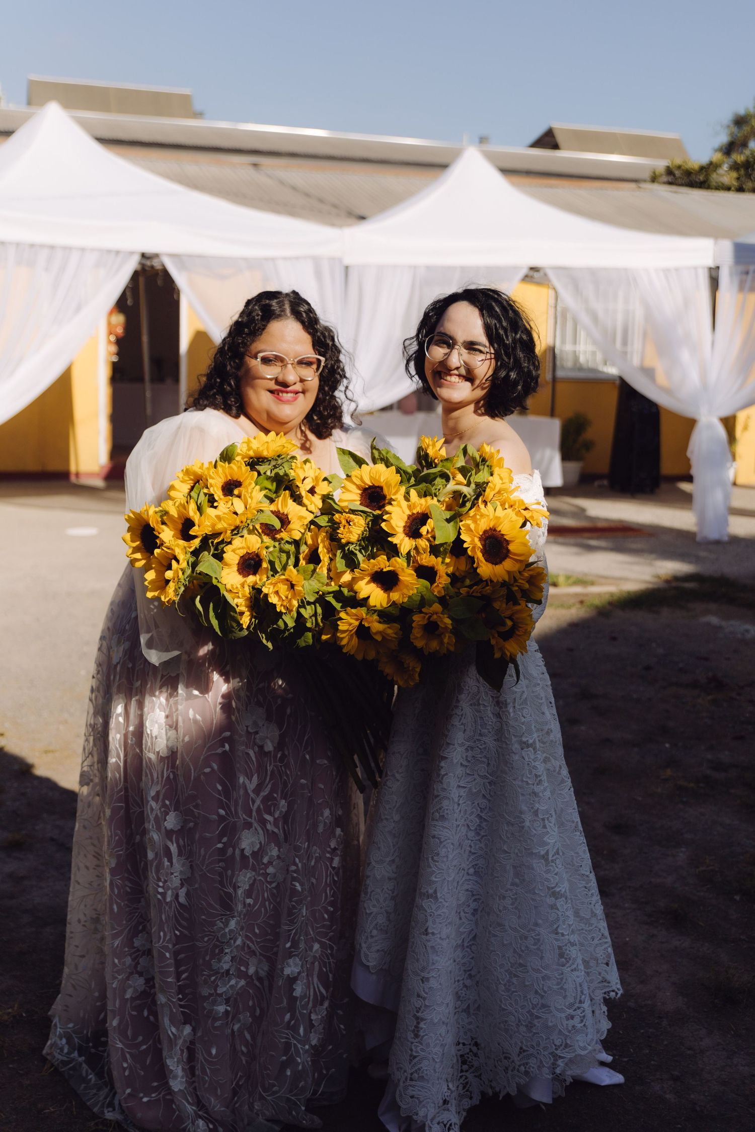 Two smiling women in lace gowns holding vibrant sunflower bouquet at outdoor wedding venue.