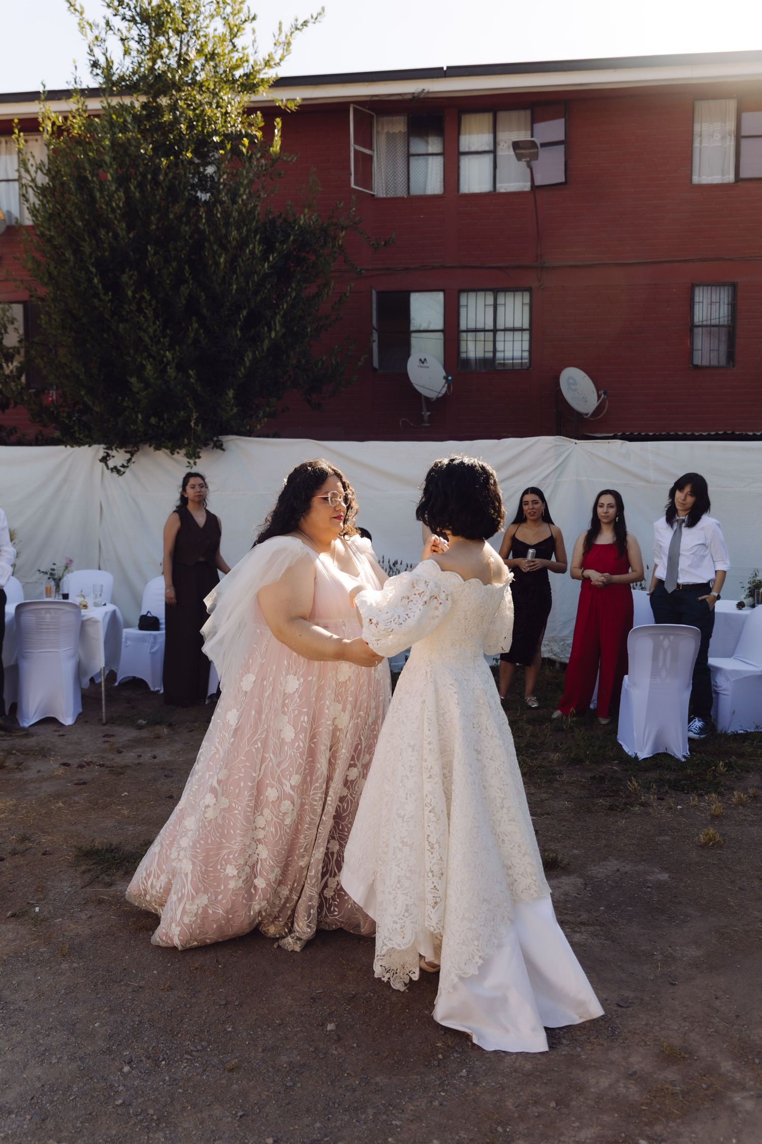Two brides sharing first dance outdoors at wedding reception with guests watching in background.