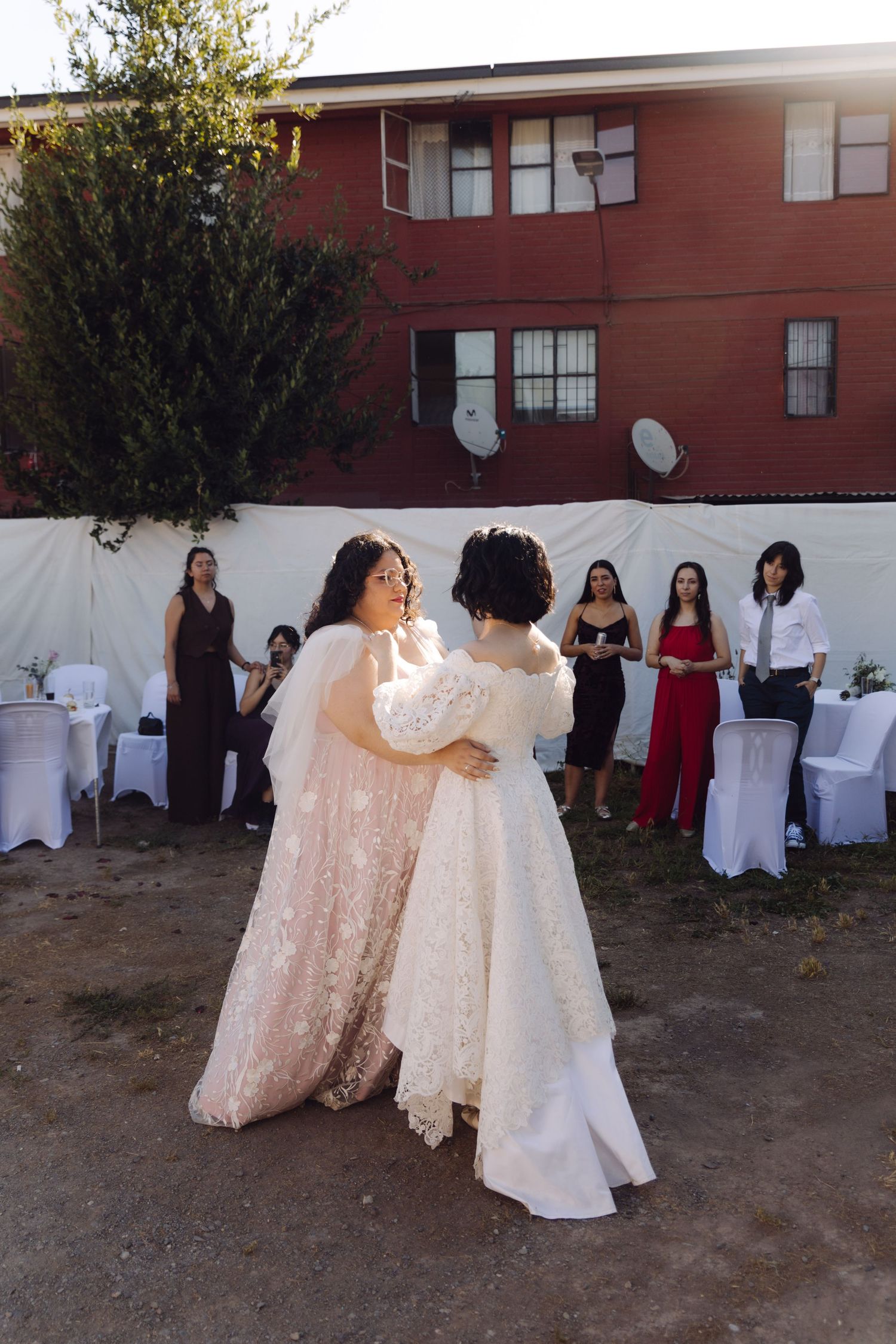 Two brides in white and blush lace gowns dancing at outdoor wedding with guests watching.