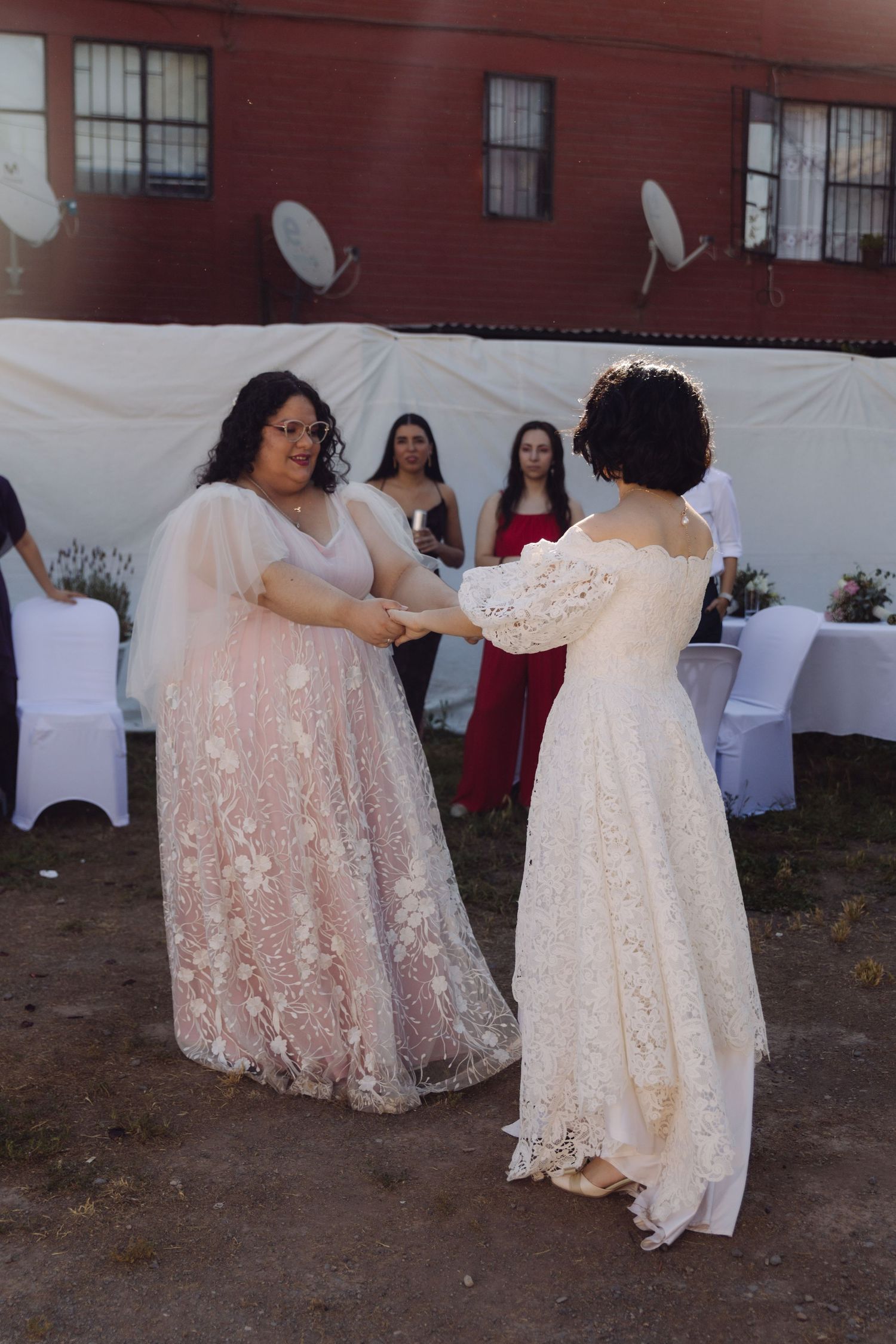 Two brides in elegant gowns dance together at outdoor wedding reception with guests watching.