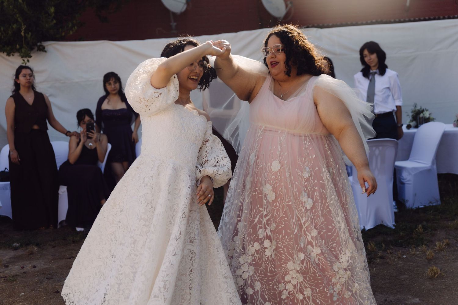 Two brides laugh joyfully while sharing first dance at outdoor wedding celebration.