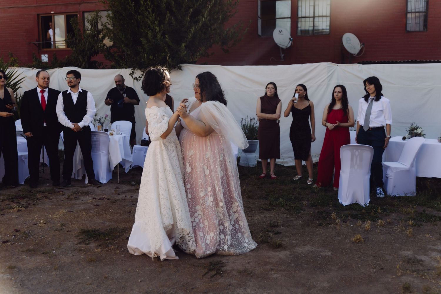 Two brides share first dance outdoors as wedding guests watch in background.