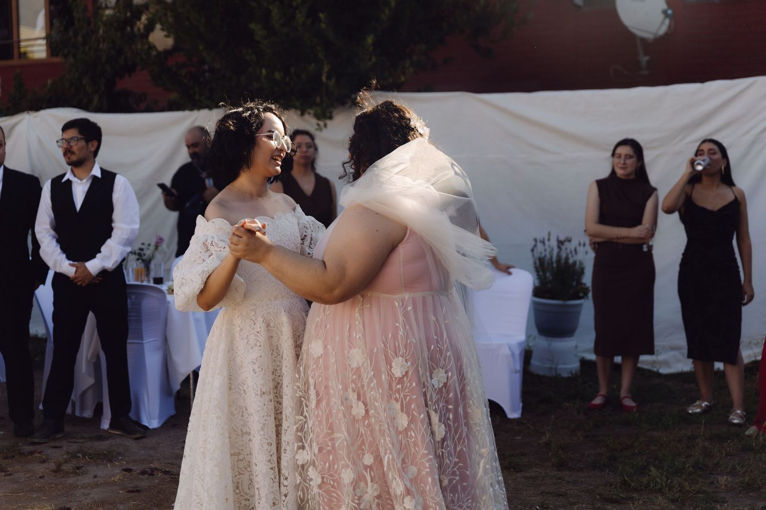 Two brides in lace and pink gowns share joyful first dance at outdoor reception.