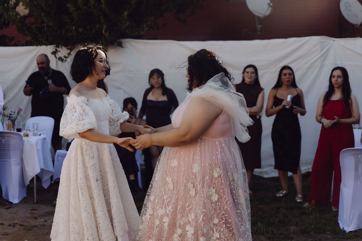 Two brides in lace wedding gowns hold hands during outdoor wedding ceremony while guests watch.