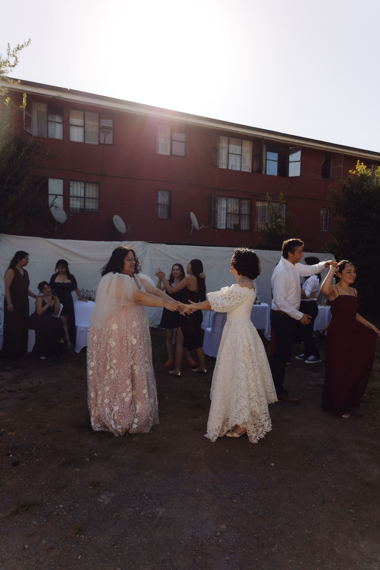 Two brides share first dance at outdoor wedding reception near red brick building at sunset.