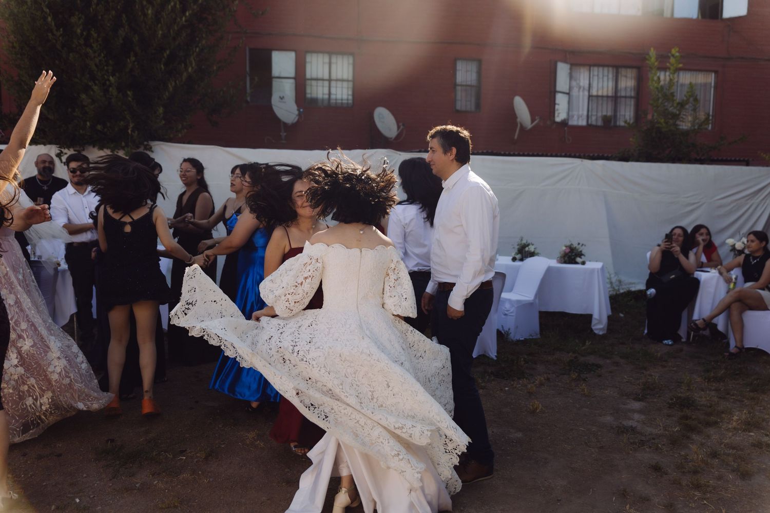 Bride in white lace wedding dress dances energetically with guests at outdoor wedding reception.