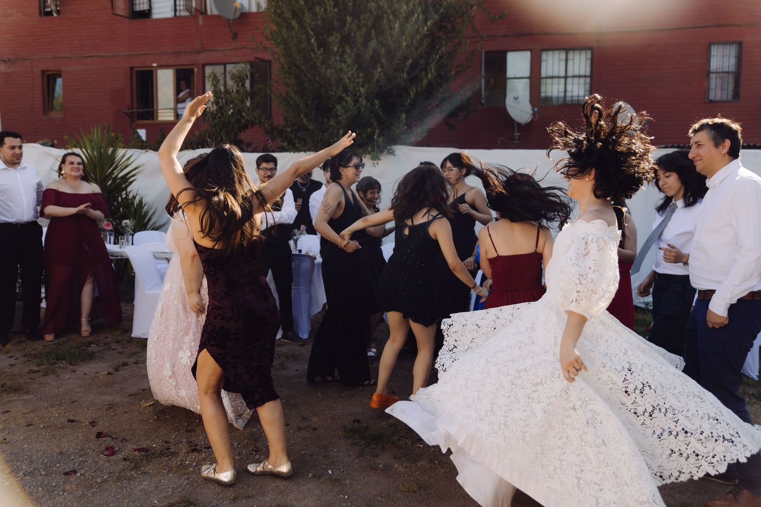 Wedding guests and bride in white lace gown dance joyfully at outdoor reception party.