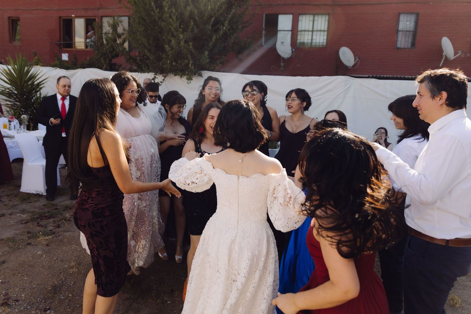 Wedding guests surround bride in white gown during celebratory outdoor reception dance.