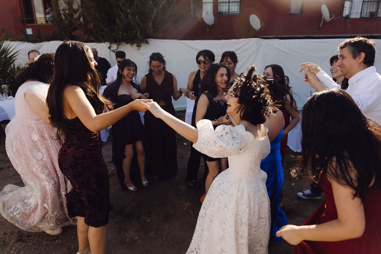 Bride in white lace dress dances with guests in circle at outdoor wedding reception.