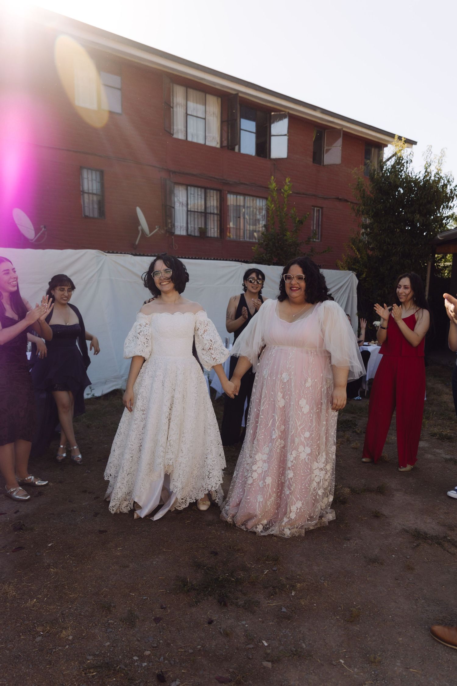Two brides in white and blush wedding gowns hold hands walking outdoors at their wedding celebration.