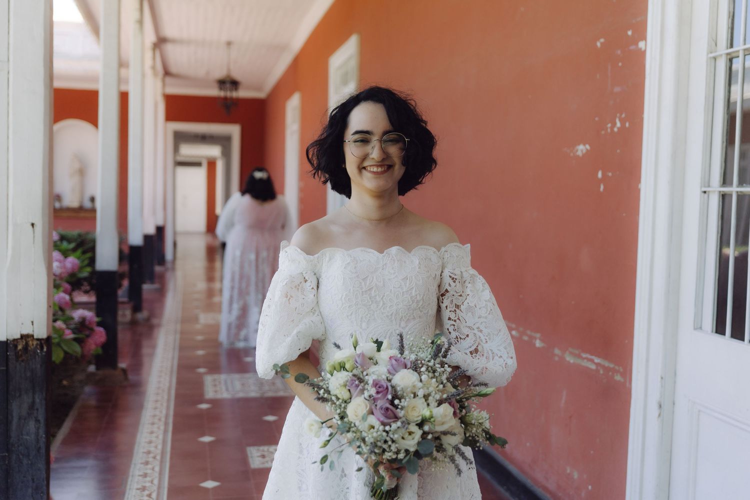 Smiling bride in off-shoulder lace gown holds purple and white bouquet in a pink hallway.