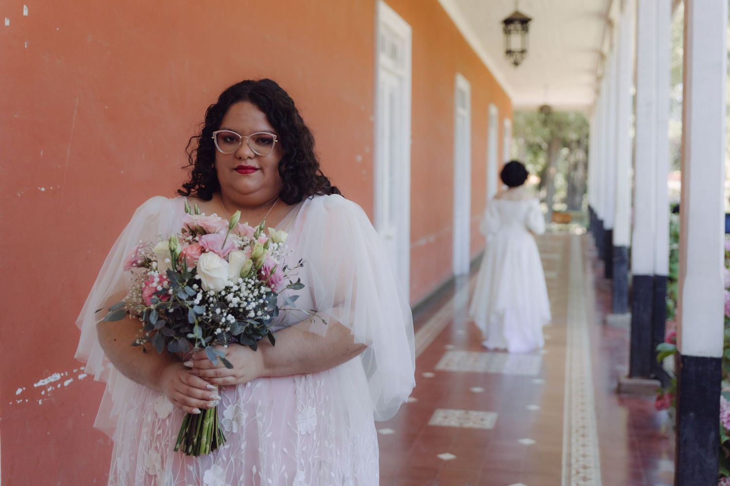 Plus-size bride in lace wedding dress holds pink floral bouquet in a coral-walled venue corridor.