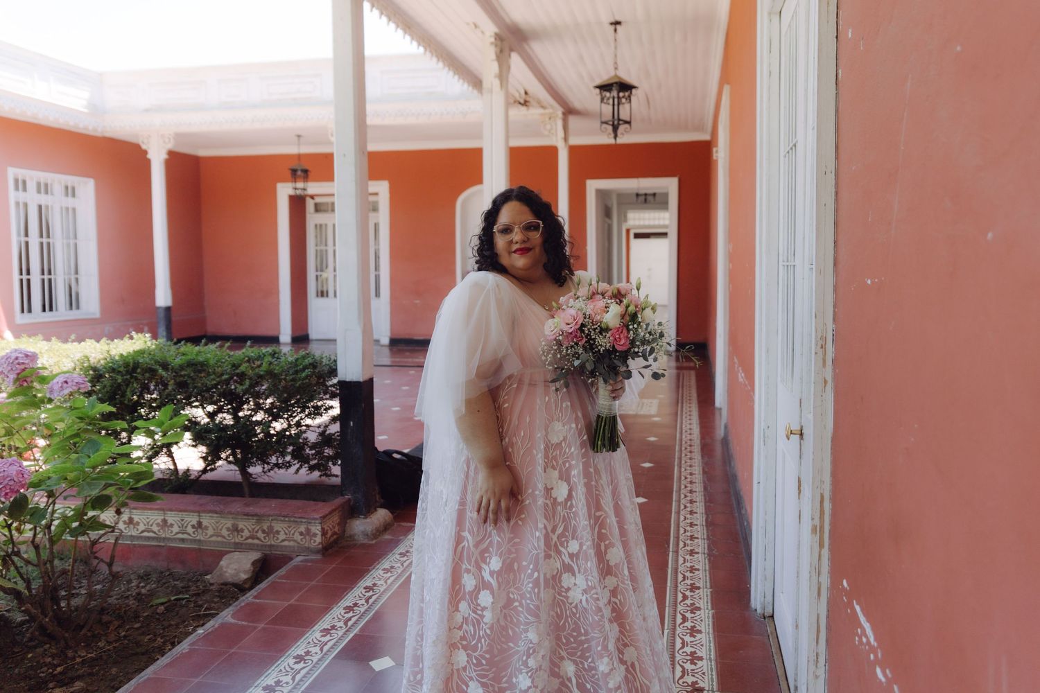 Bride in blush lace wedding gown with veil poses with floral bouquet on a pink building porch.