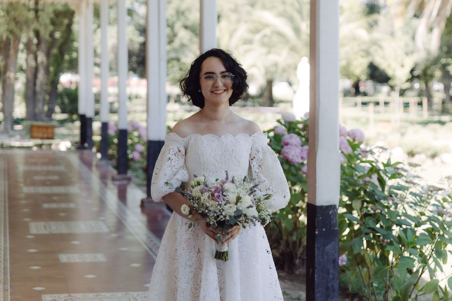 Bride in off-shoulder white lace dress smiles holding bouquet in a garden colonnade.