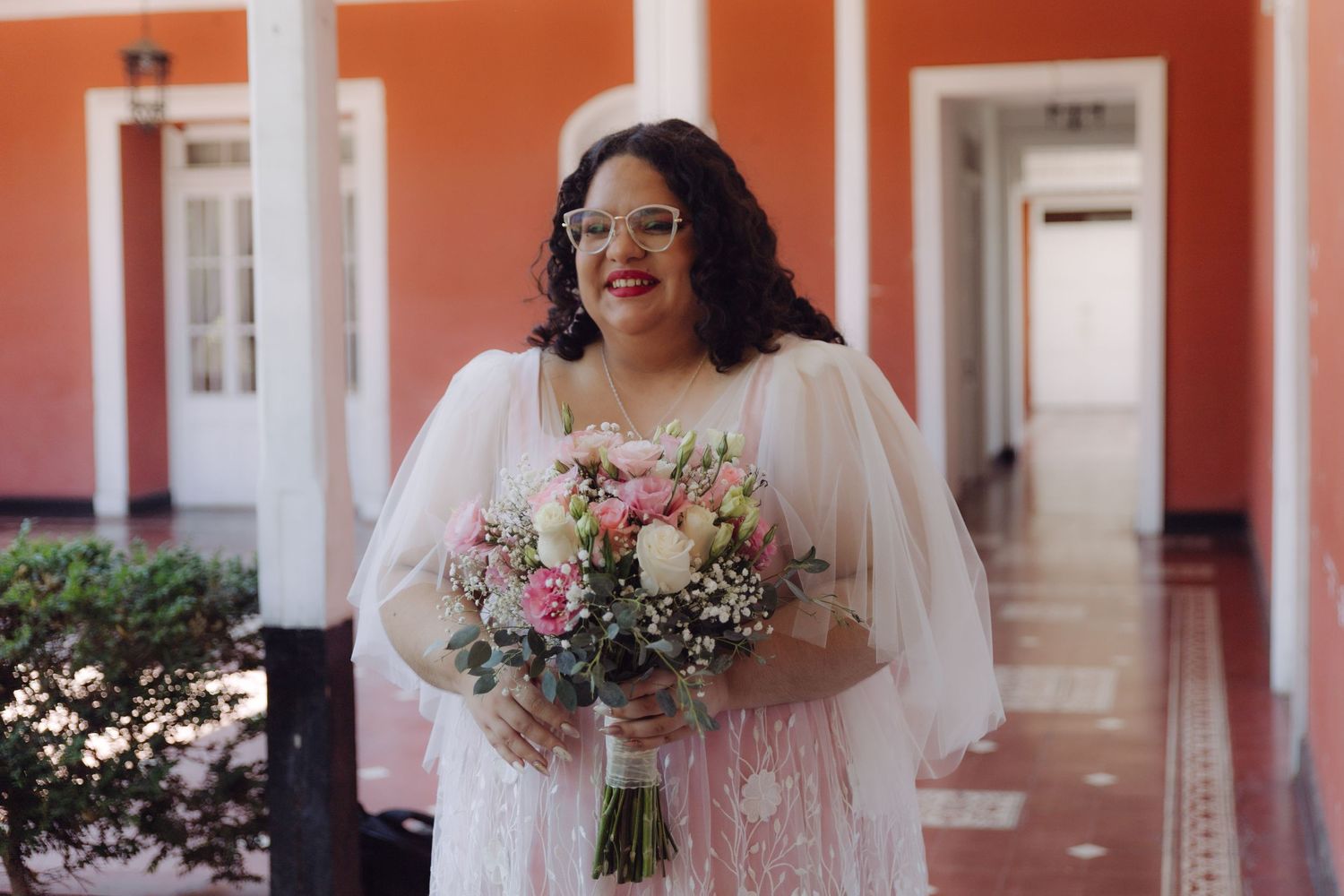 Smiling bride in white lace gown with veil holds pink rose bouquet in a coral archway corridor.