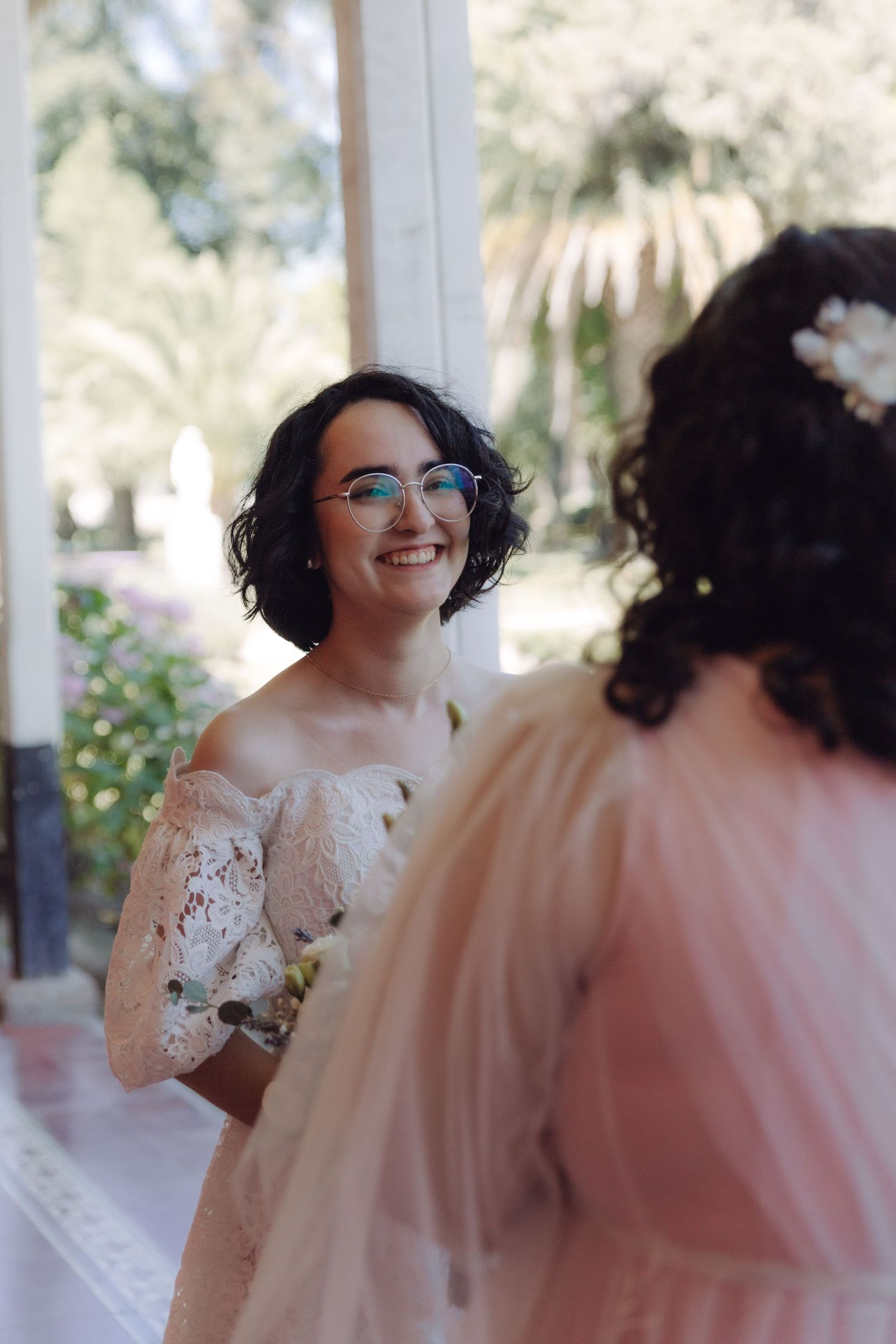 Smiling bride with glasses in off-shoulder lace gown faces another bride in blush dress outdoors.