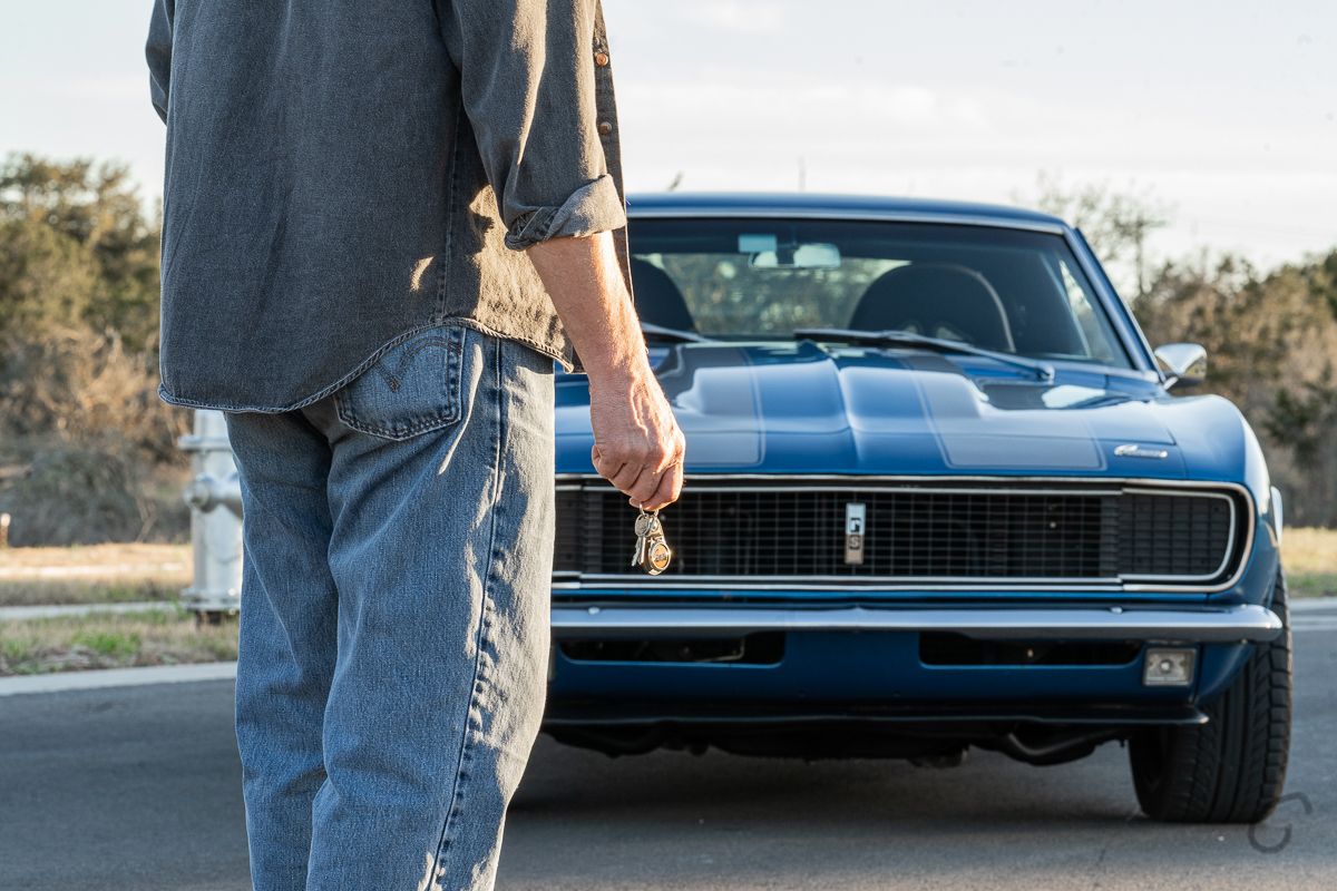 Man holding car keys standing in front of a classic blue Chevrolet Camaro muscle car on an open road.