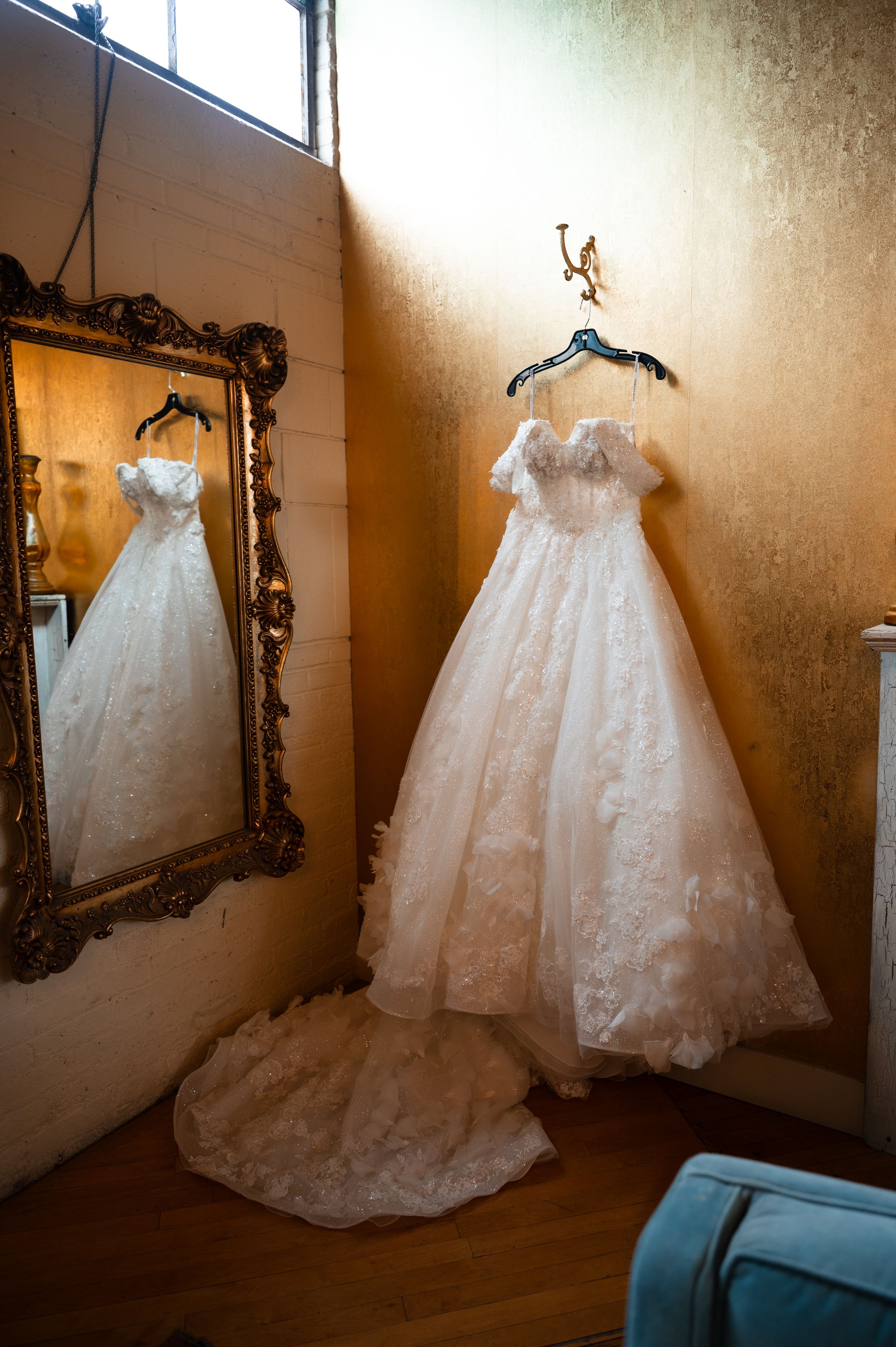 Elegant white lace ball gown wedding dress hanging on wall beside ornate gold mirror in rustic venue.