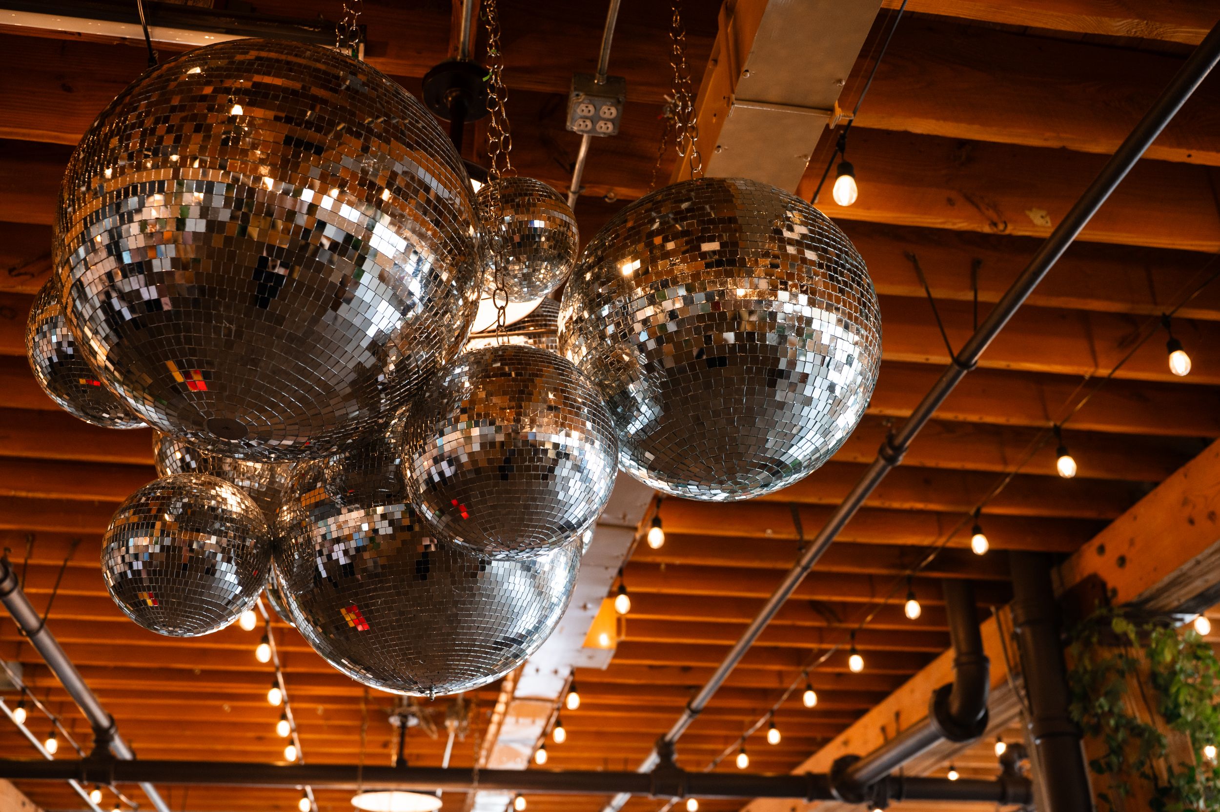Cluster of silver mirror disco balls hanging from wooden ceiling with string lights at wedding venue.