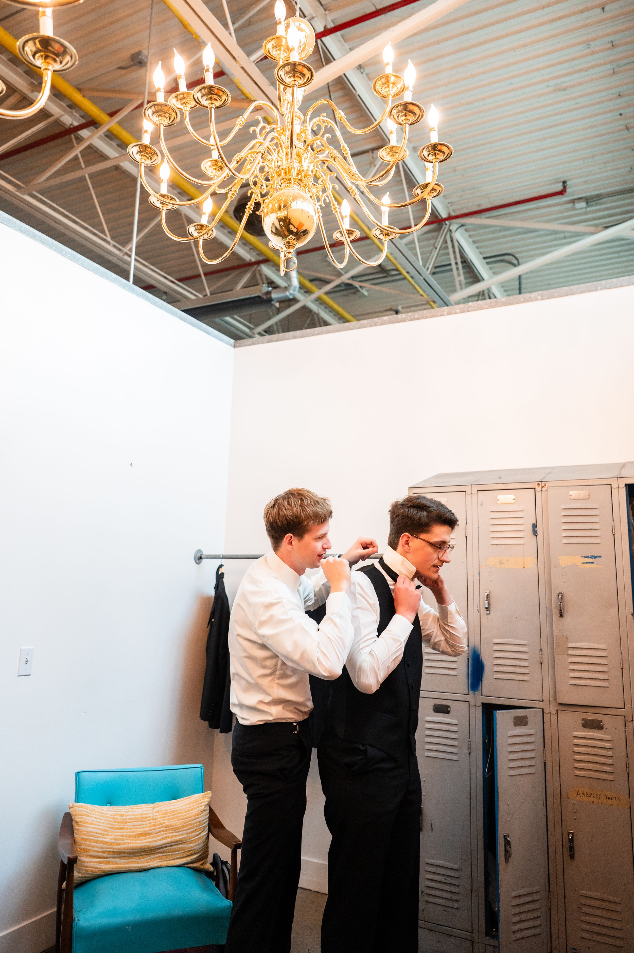 Two groomsmen in white shirts and black vests helping each other with ties near lockers under a gold chandelier.